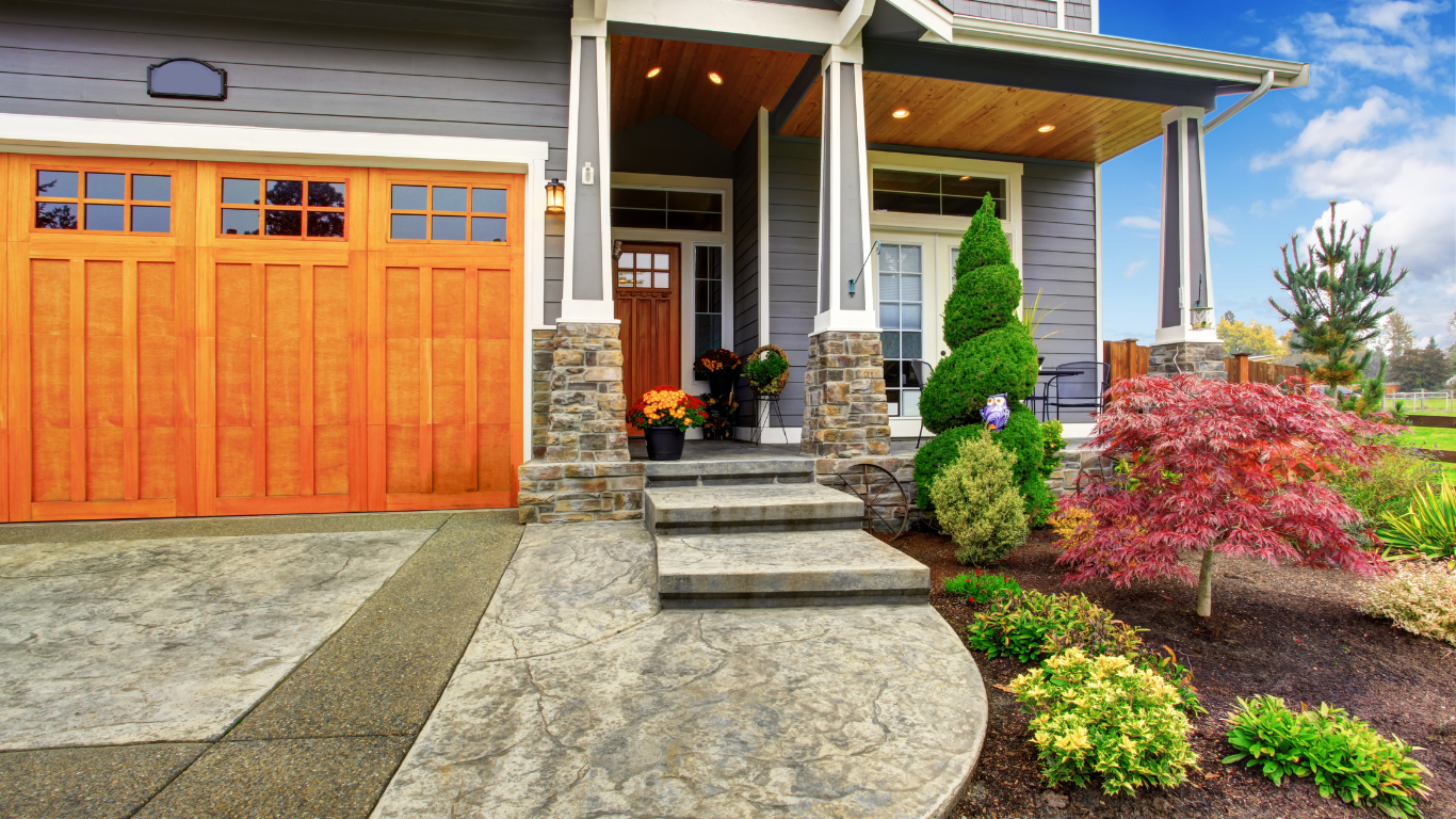 Gray house with orange garage door and entryway, stone columns, and landscaped front yard with colorful plants.