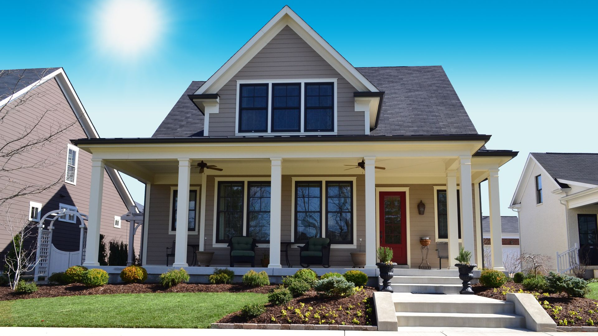 Tan house with porch, front steps, and black roof under a blue sky with bright sun.