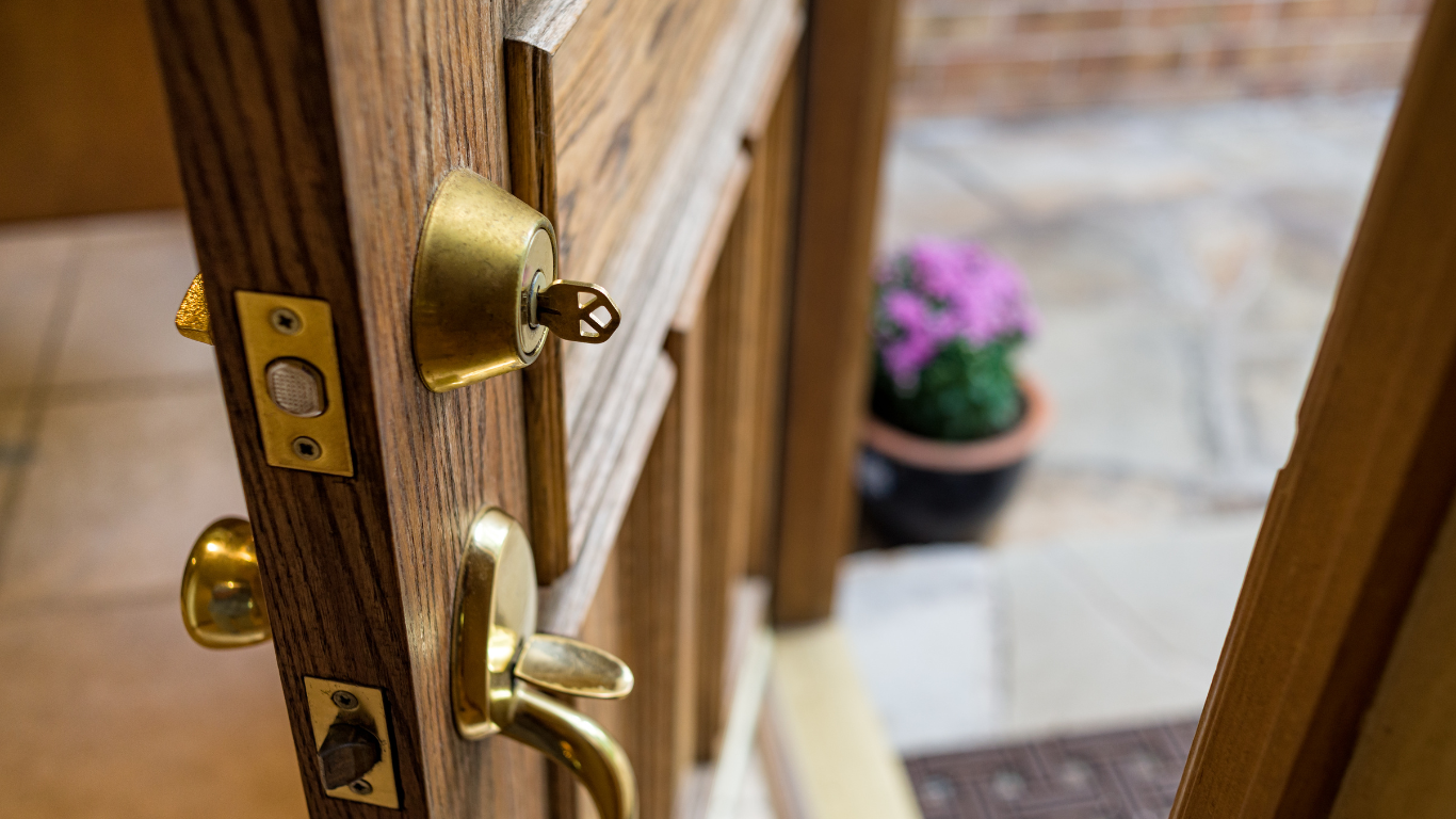 Close-up of a wooden door, partially open, with a key in the lock; a potted plant is visible outside.