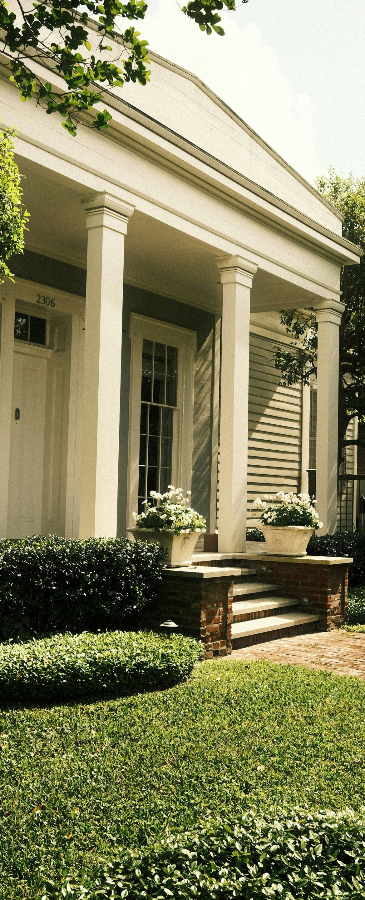 A cream-colored cottage with a front porch, tall white columns, a triangular pediment, and a lush green lawn.