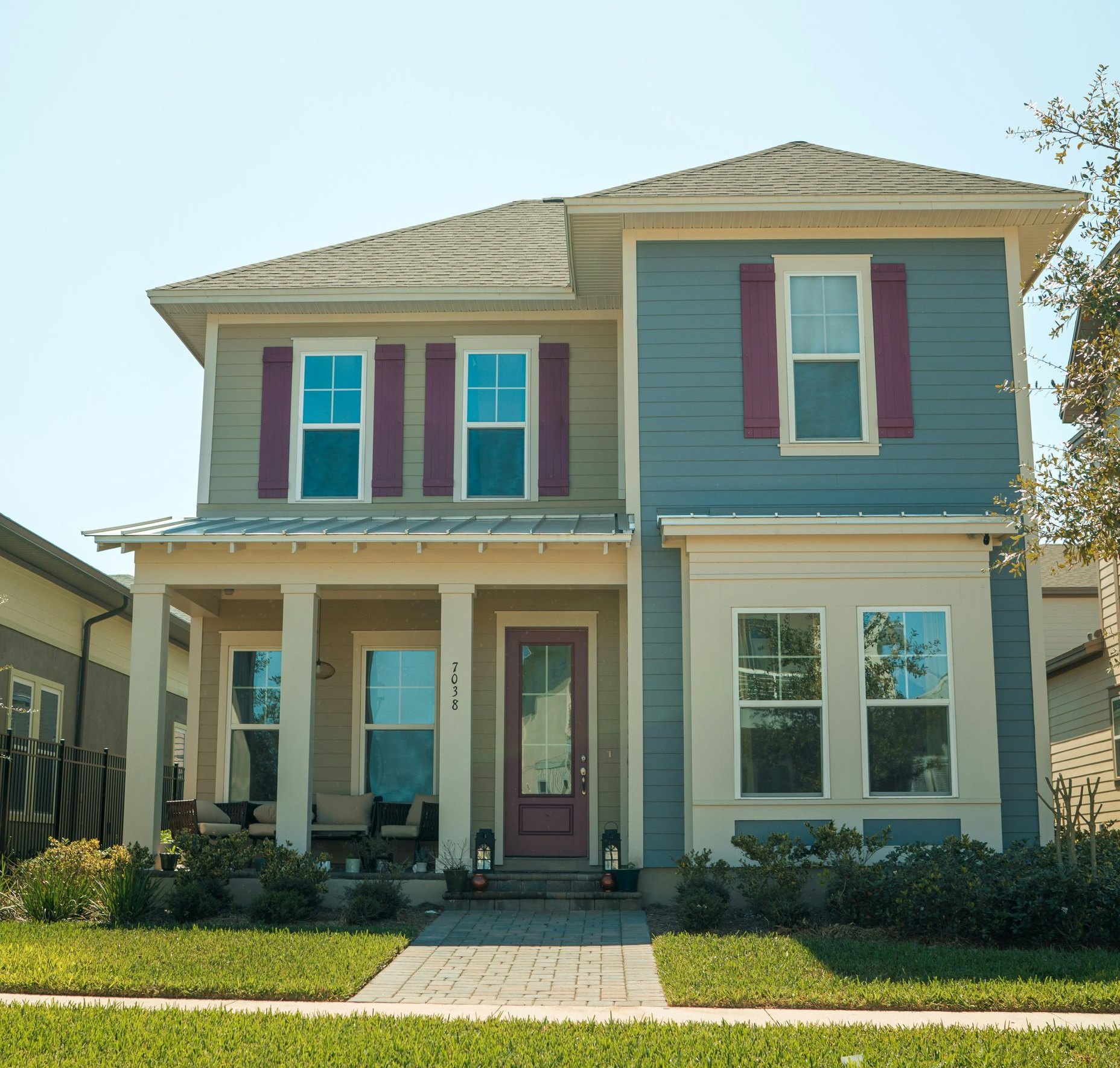 Two-story house with green lawn, blue siding, red shutters, and a front porch under a sunny sky.