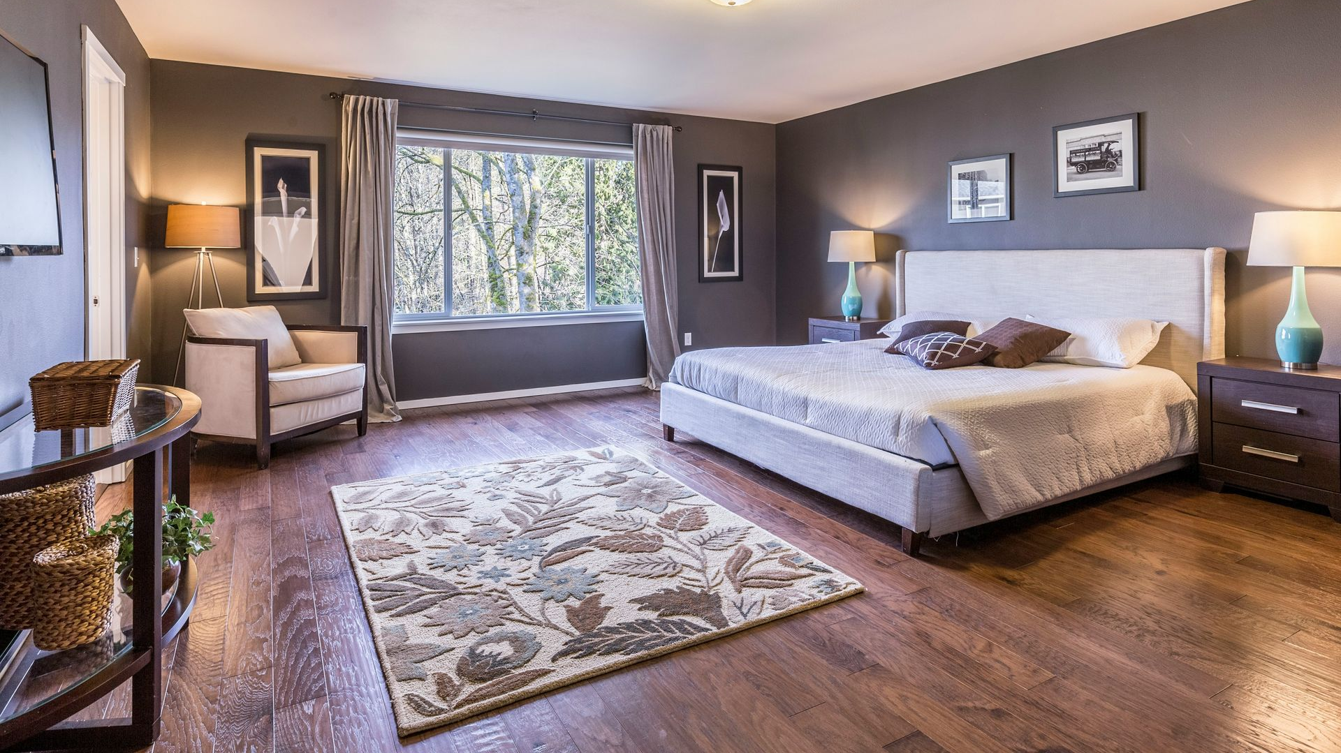 Bedroom with hardwood floors, dark gray walls, large window with trees, bed, and sitting area.