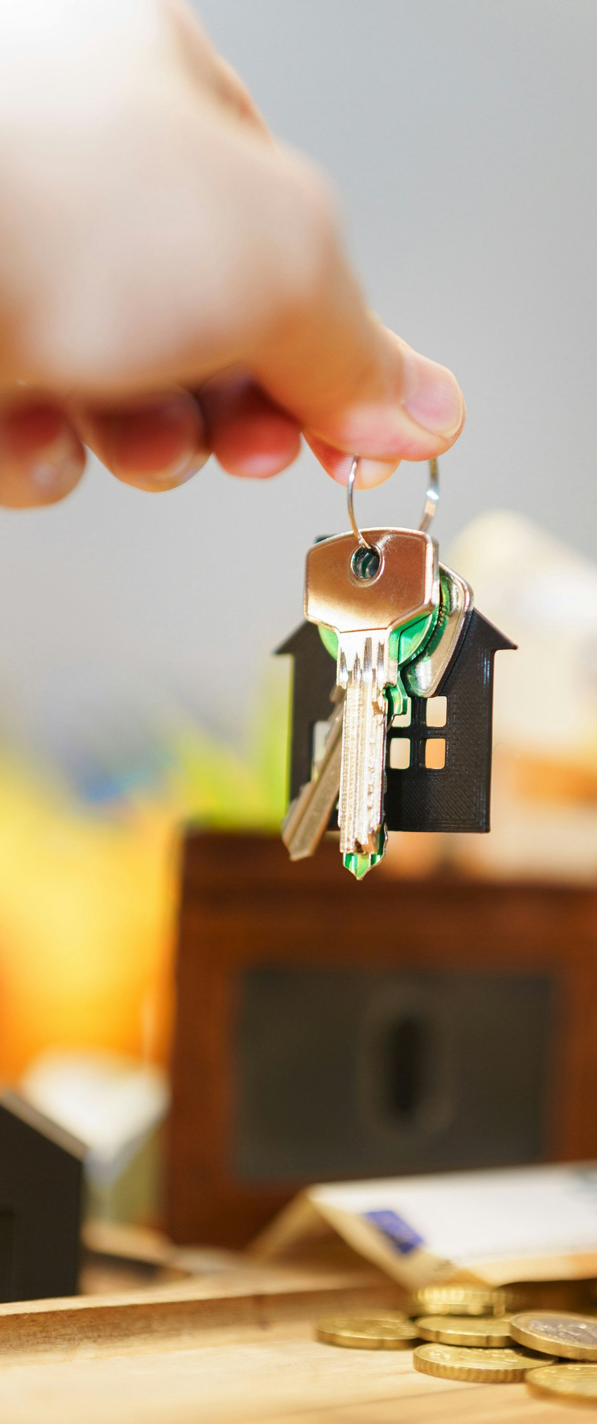 A hand holds a silver key attached to a house-shaped keychain above a brown wallet and scattered coins.