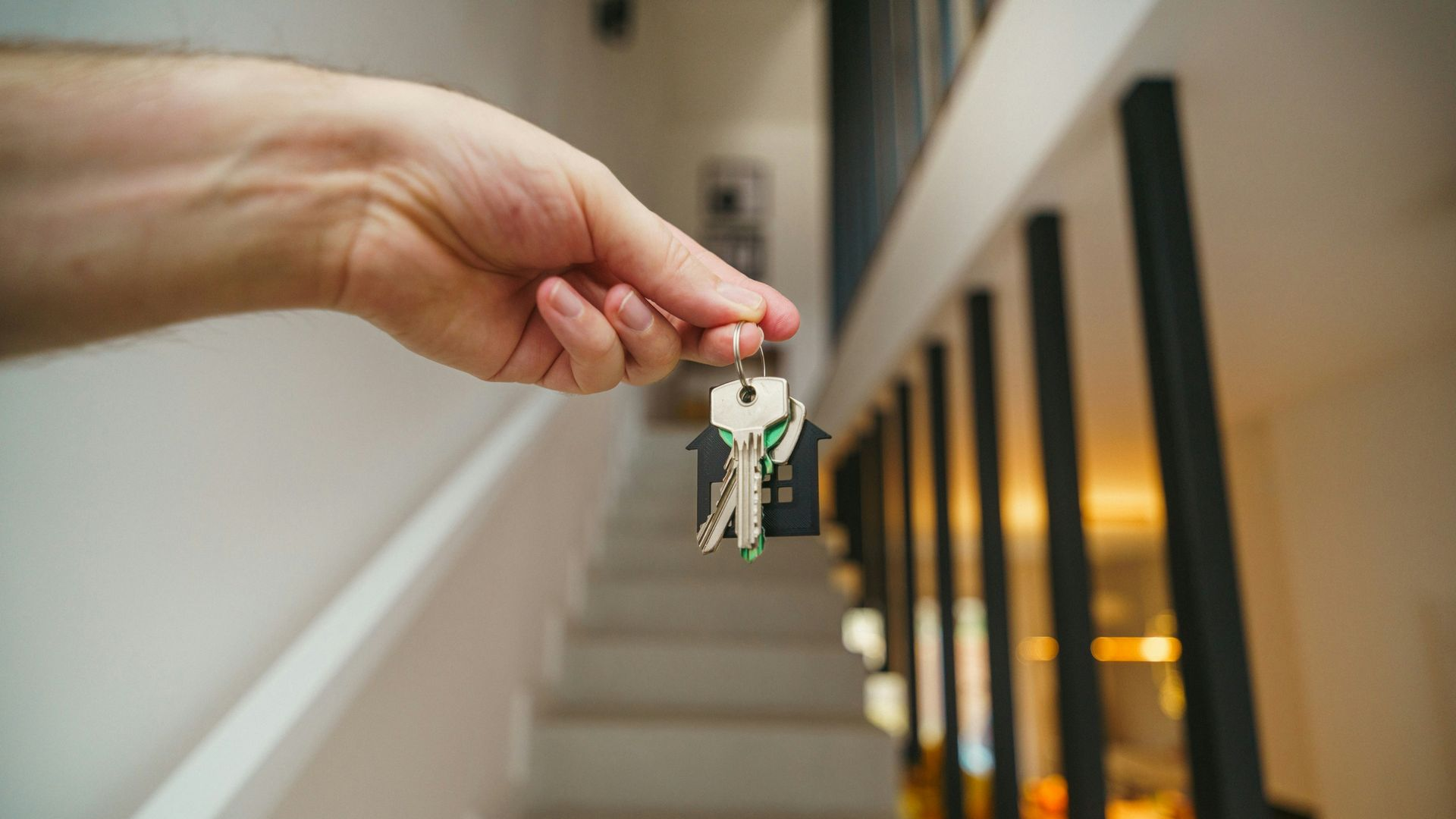 A hand holds keys with a house-shaped keychain in front of a staircase in a modern, light-filled interior.