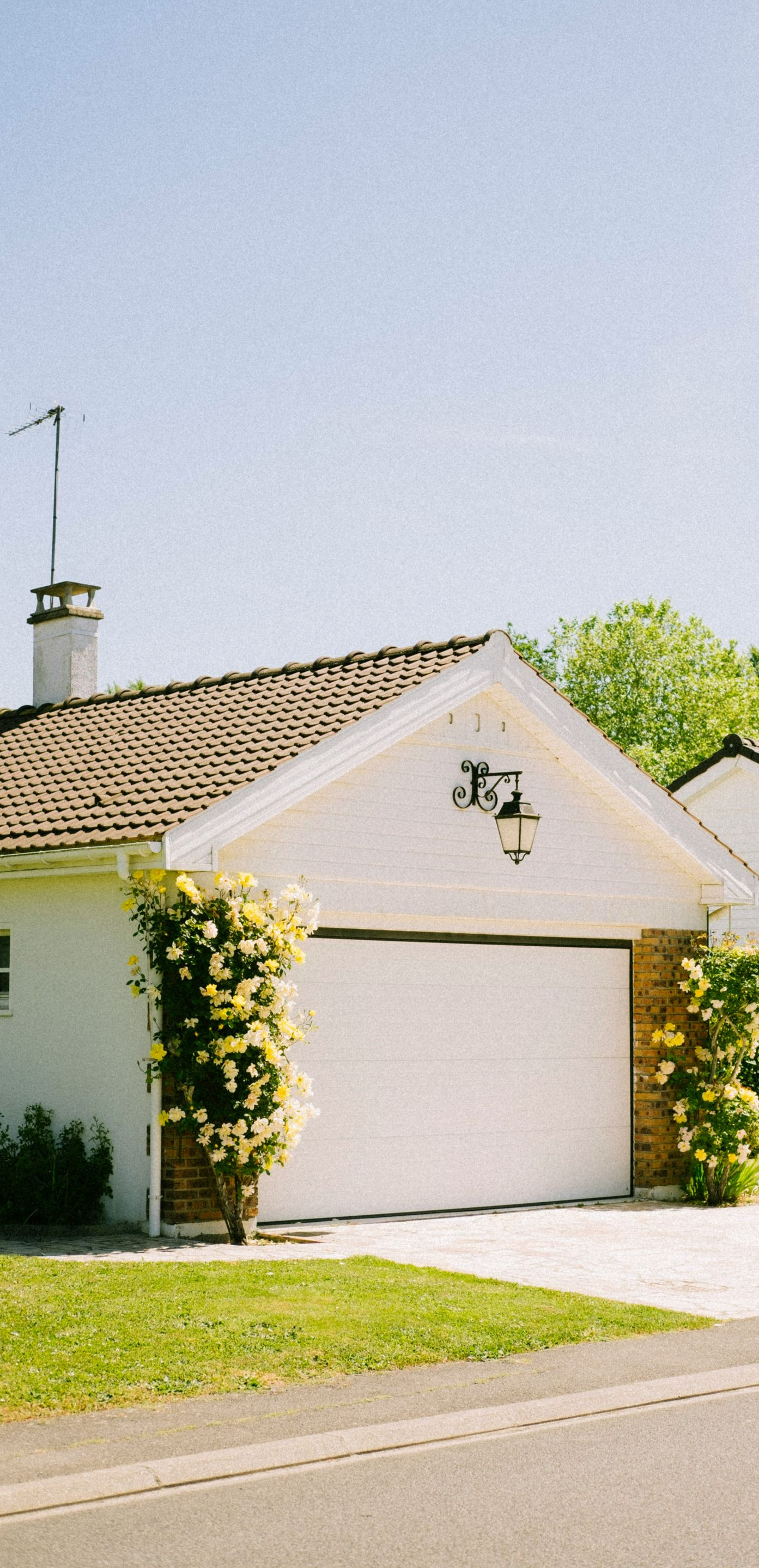 White garage with a white door, brown tiled roof, and yellow climbing roses. A chimney and an antenna are visible.