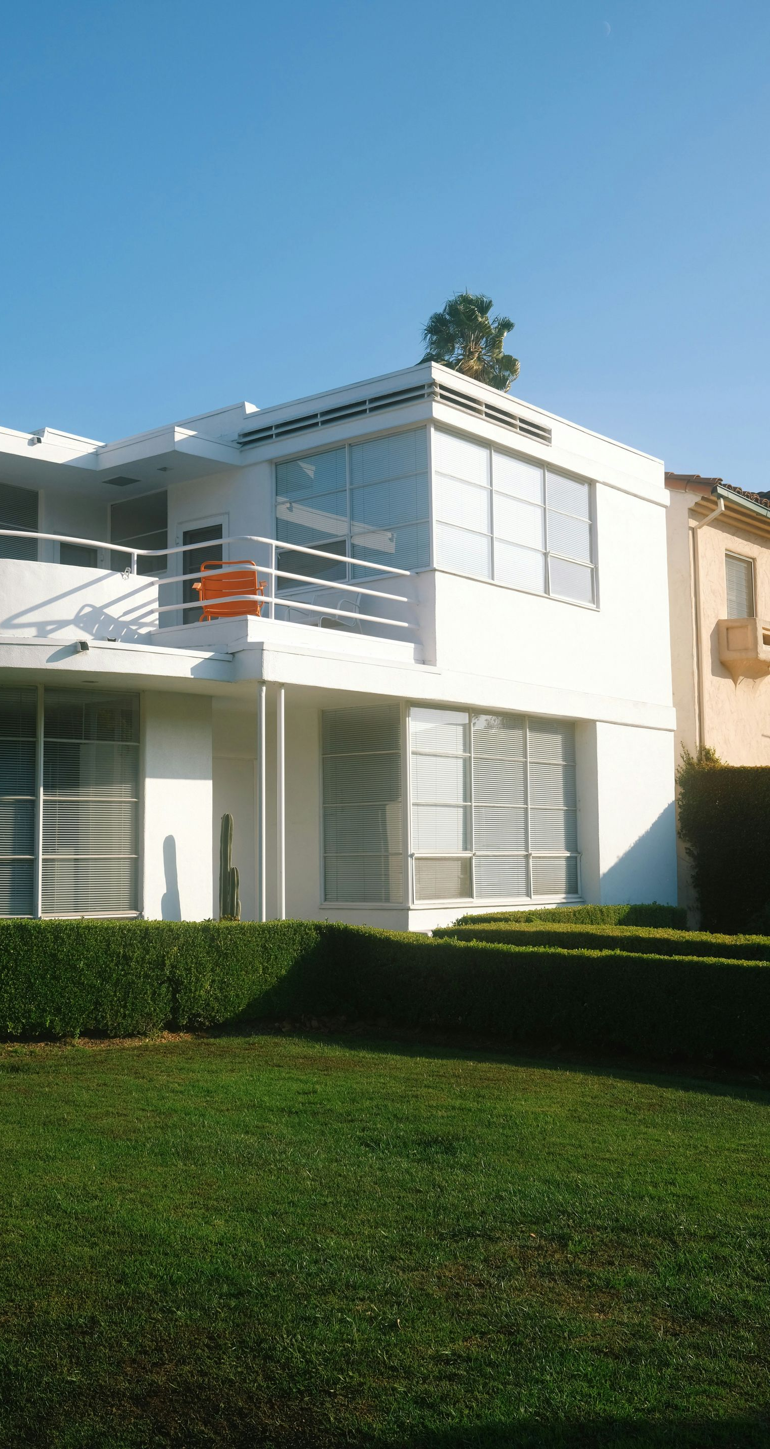 White modern two-story house with large windows, balcony, and manicured hedge on a sunny day.