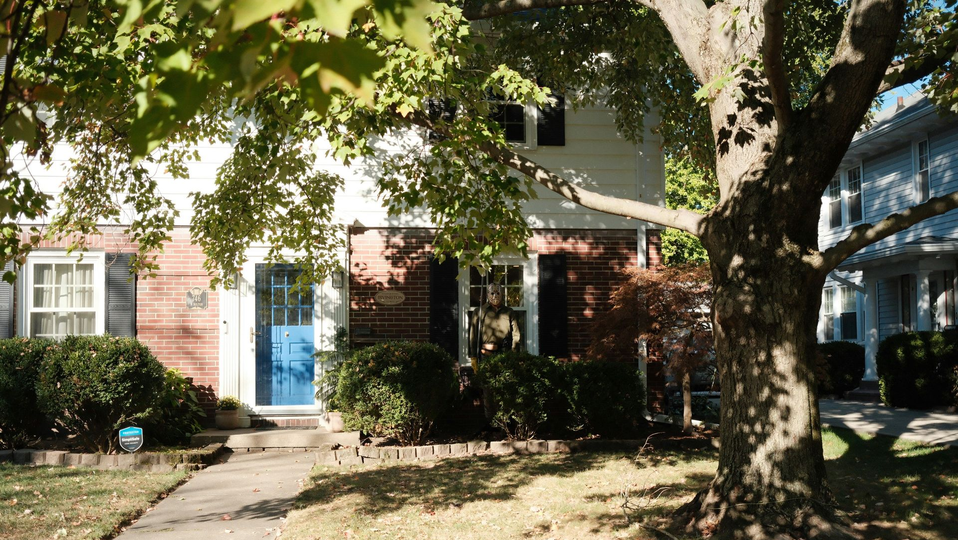 A two-story brick and white home with a blue front door, surrounded by green shrubs and a large tree in the front yard.