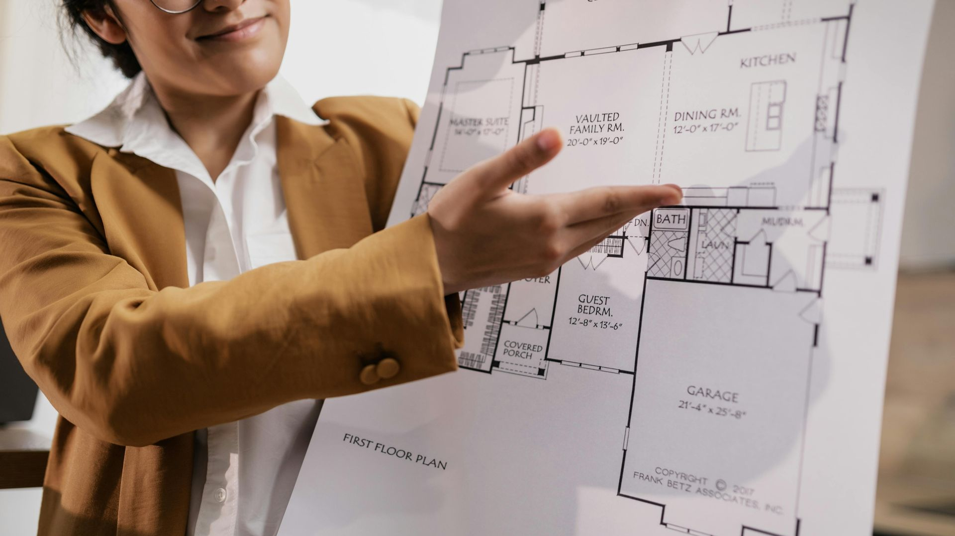 Woman in a blazer pointing at a house floor plan, showing layout details.