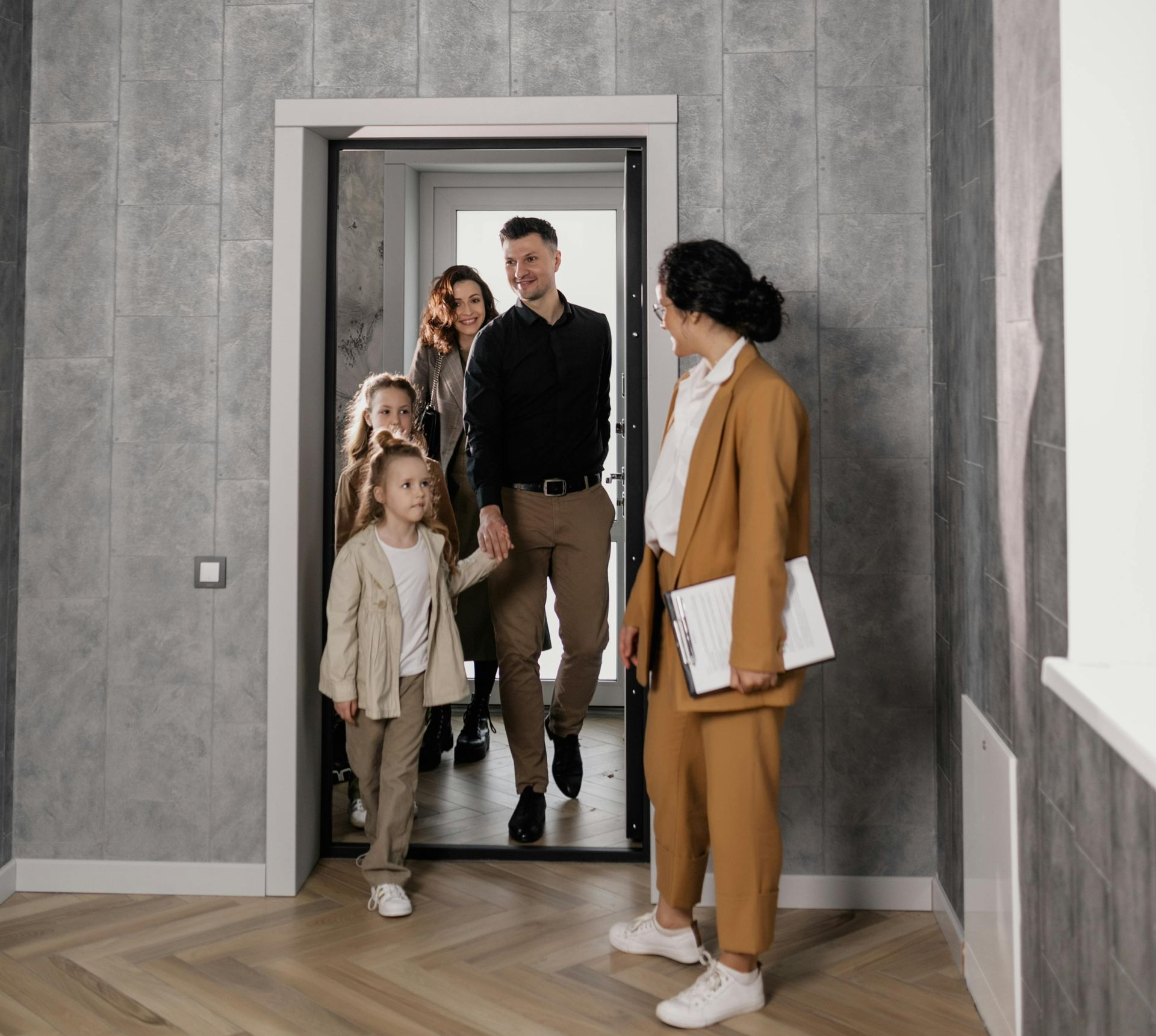 Real estate agent in a brown suit greets a family exiting a doorway.