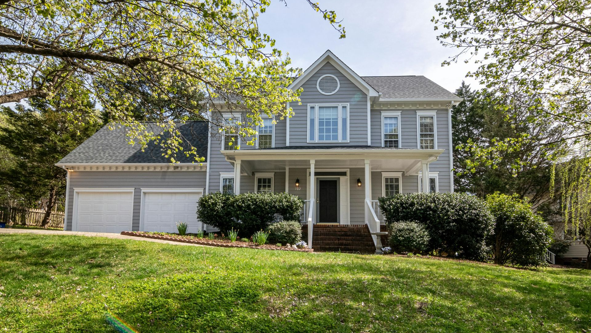 Gray two-story house with white trim, a front porch, and a two-car garage, on a green lawn with trees.