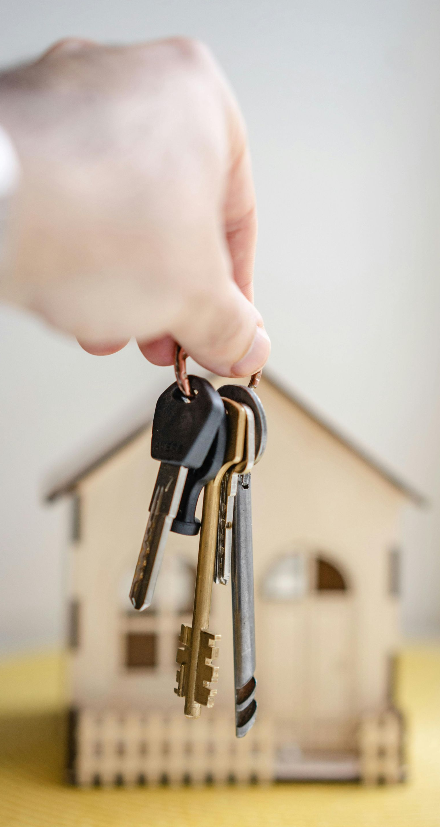 Hand holding a set of keys in front of a small wooden house cutout.