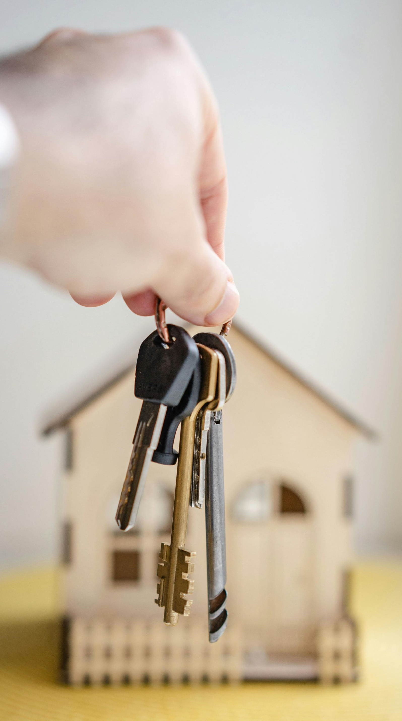 Hand holding keys in front of a miniature wooden house, suggesting homeownership.