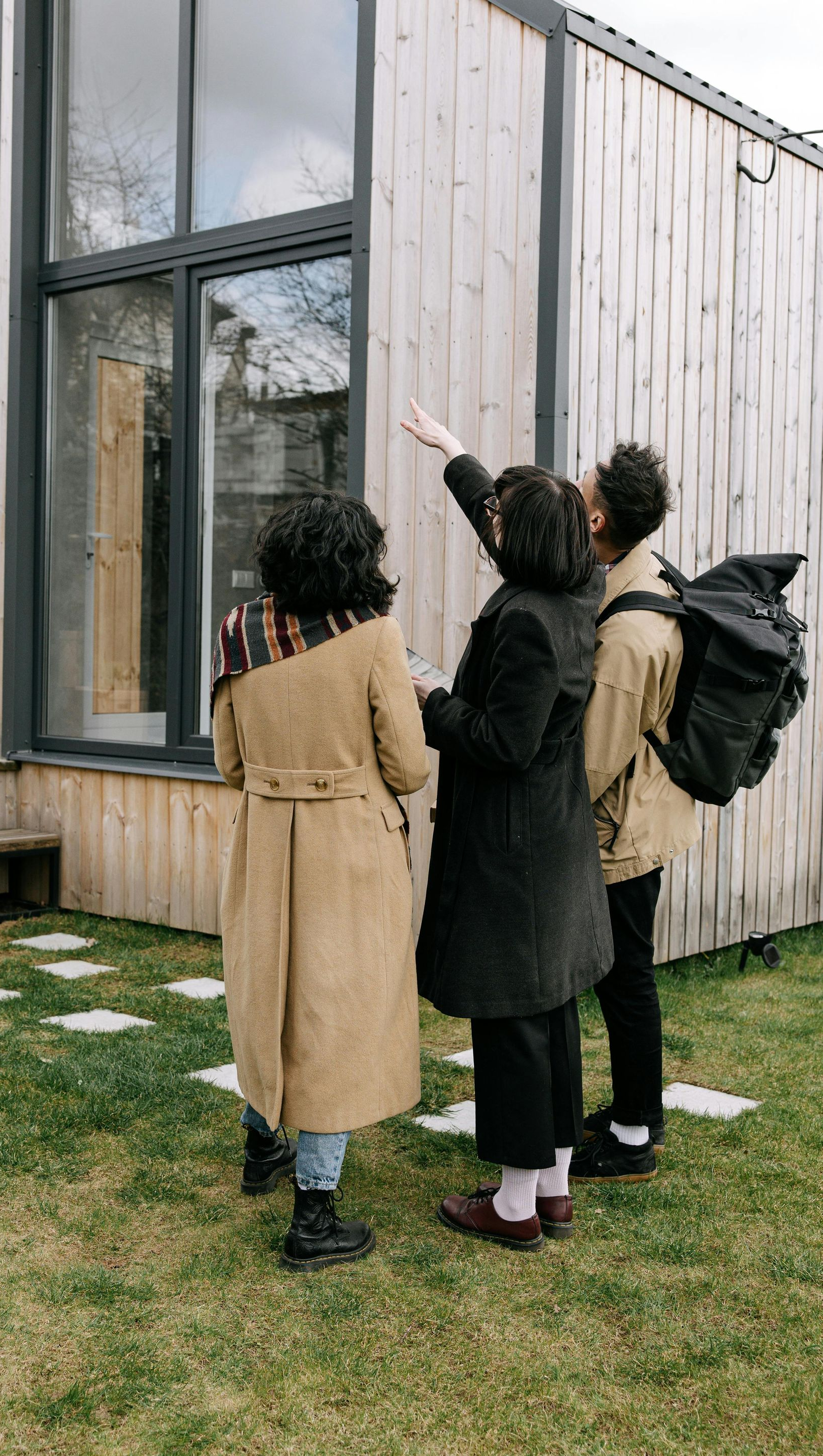 Four people looking at a modern house, one points up. Green grass, wooden exterior.
