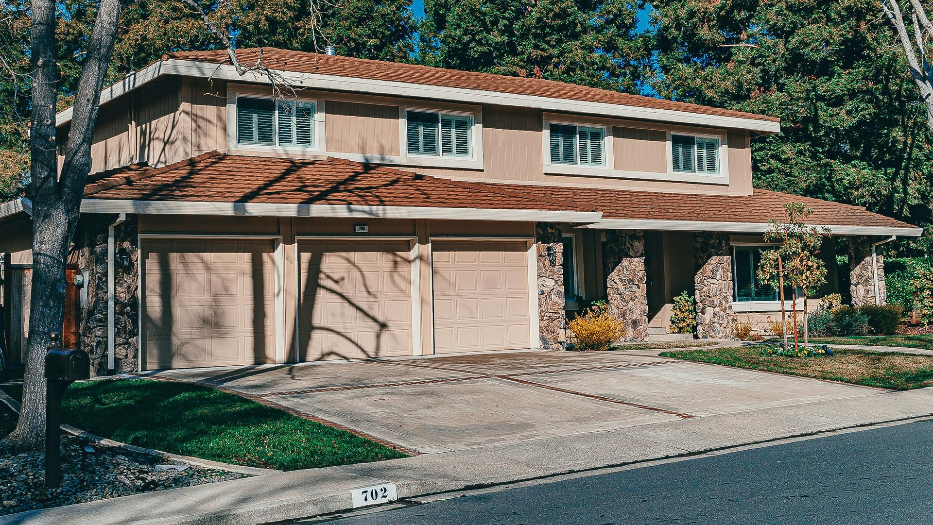 Two-story beige house with three-car garage, red tile roof, and stone accents.