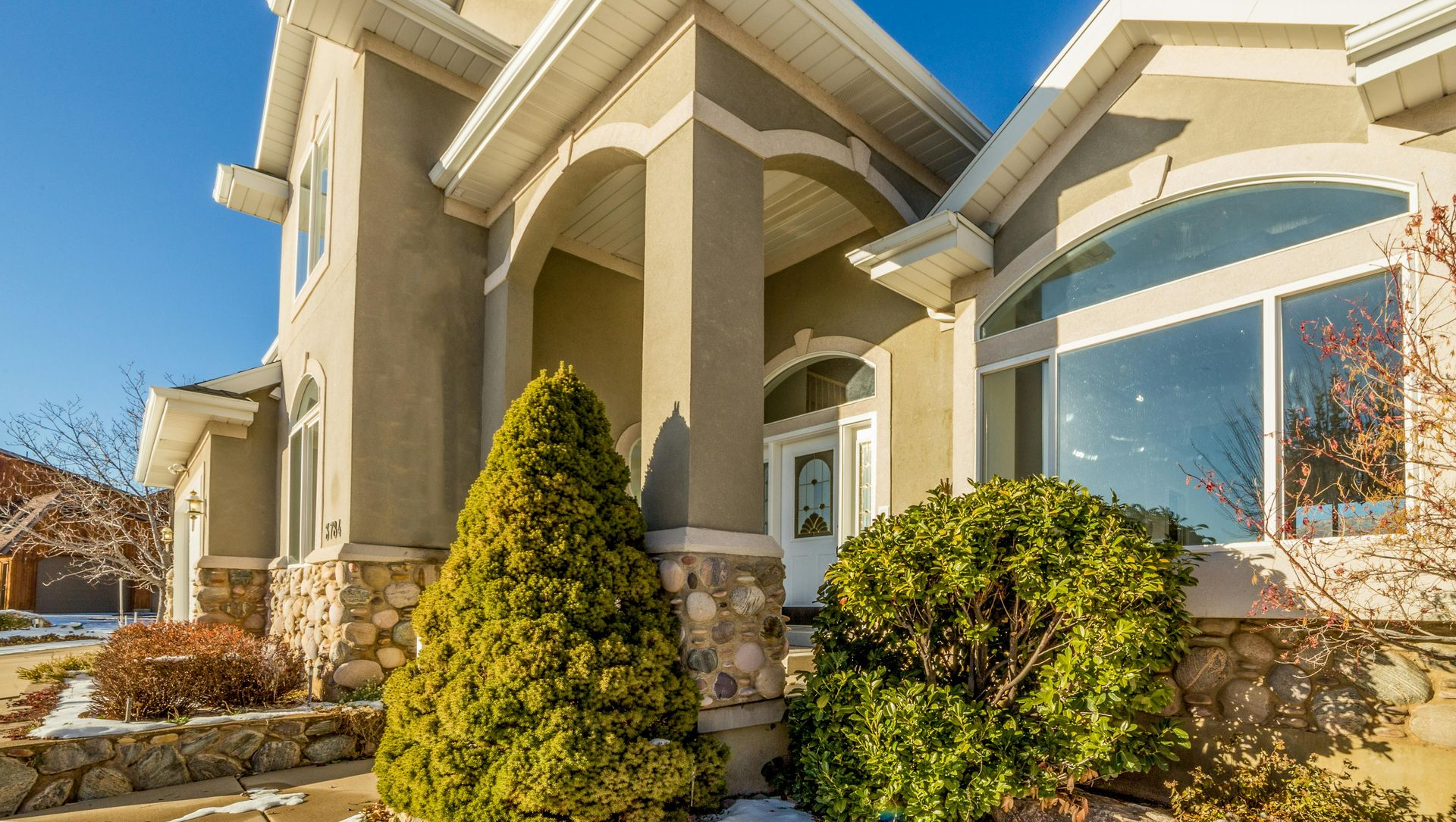Beige stucco house with stone accents, arched entryway, large windows, and manicured shrubs under a blue sky.