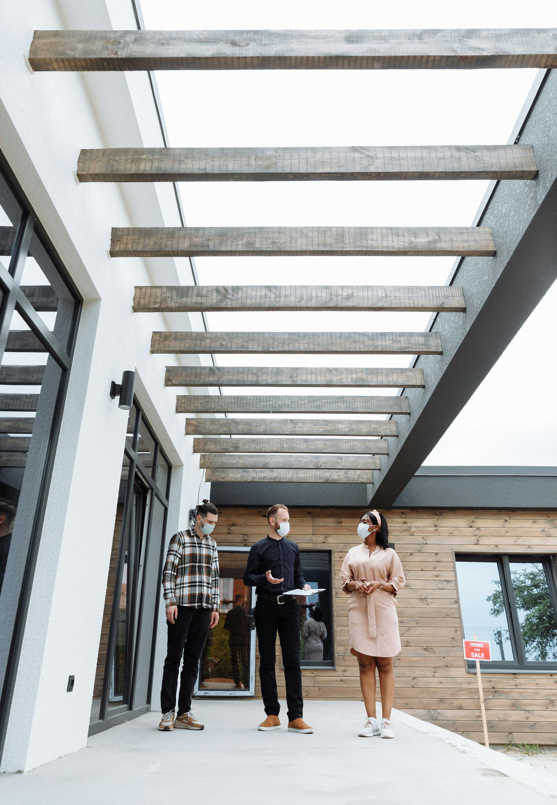 Three people wearing masks stand under a wooden pergola, looking at a modern house.