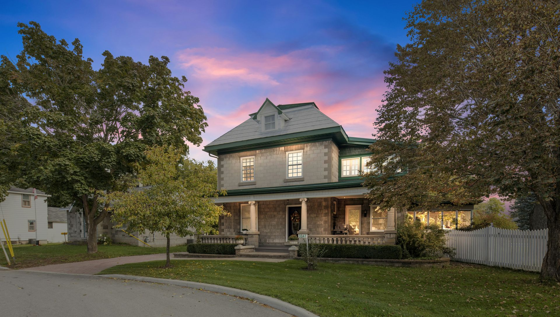 Two-story stone house with a gabled roof, porch, and mature trees, set against a twilight sky.