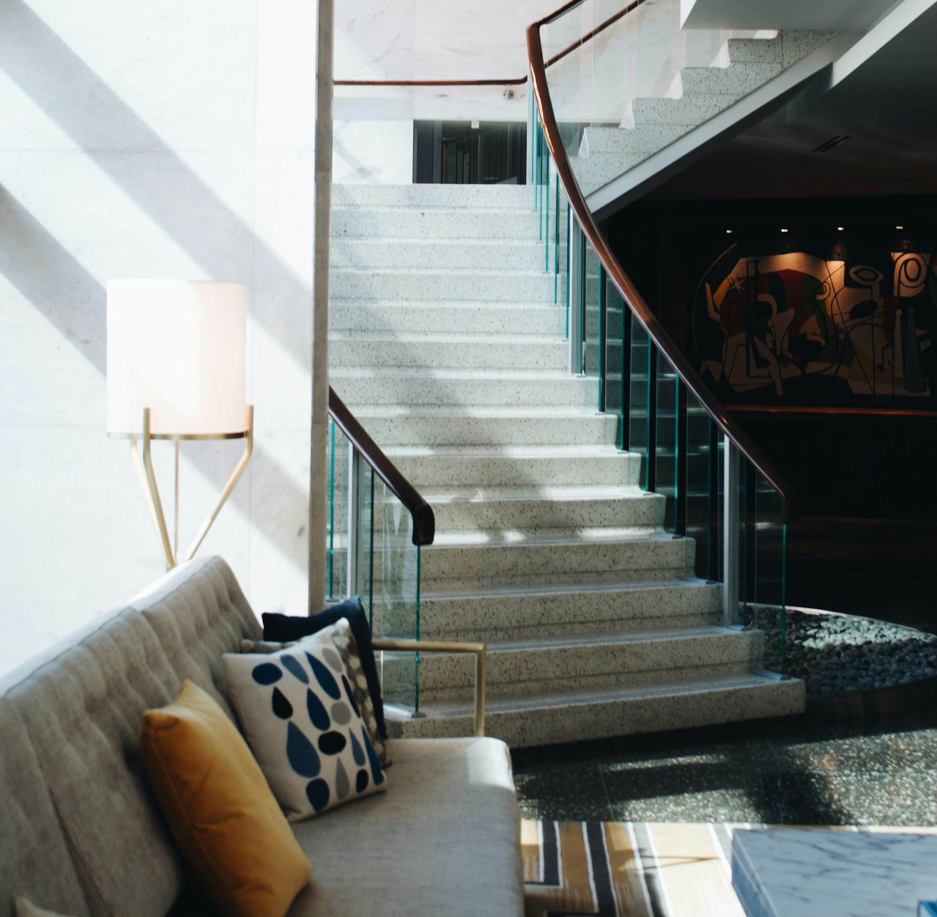 A gray couch with colorful pillows sits near a terrazzo staircase with a wooden railing; a gold lamp stands beside the couch.