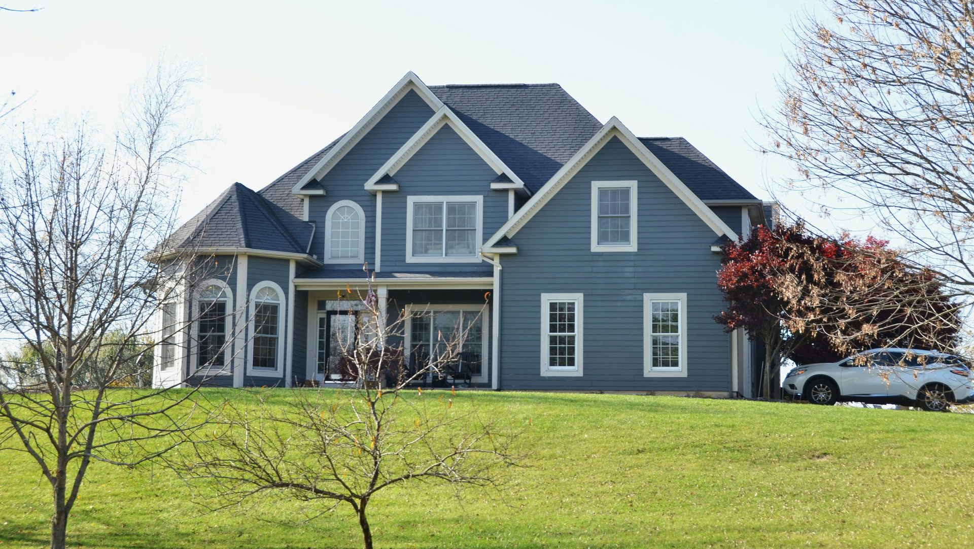 A two-story blue house with white trim and a dark roof, situated on a grassy hill with trees and a parked car.