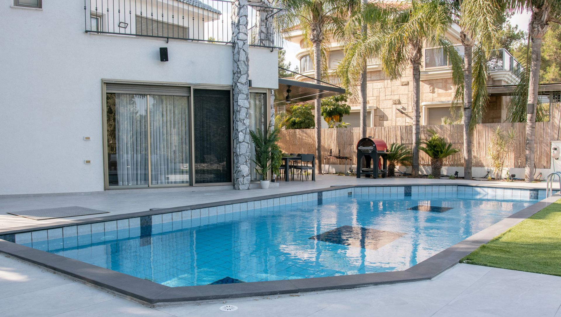 A rectangular swimming pool with blue water sits on a light gray stone patio beside a modern white house and palm trees.