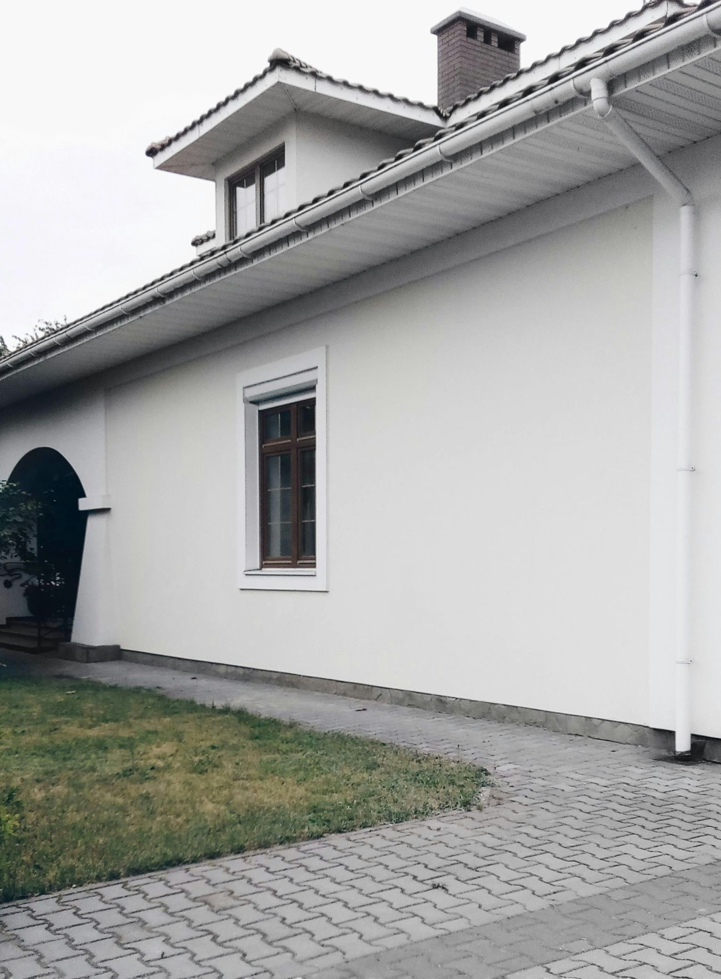 White house with a brown window, arched entryway, dormer, and gray brick driveway.