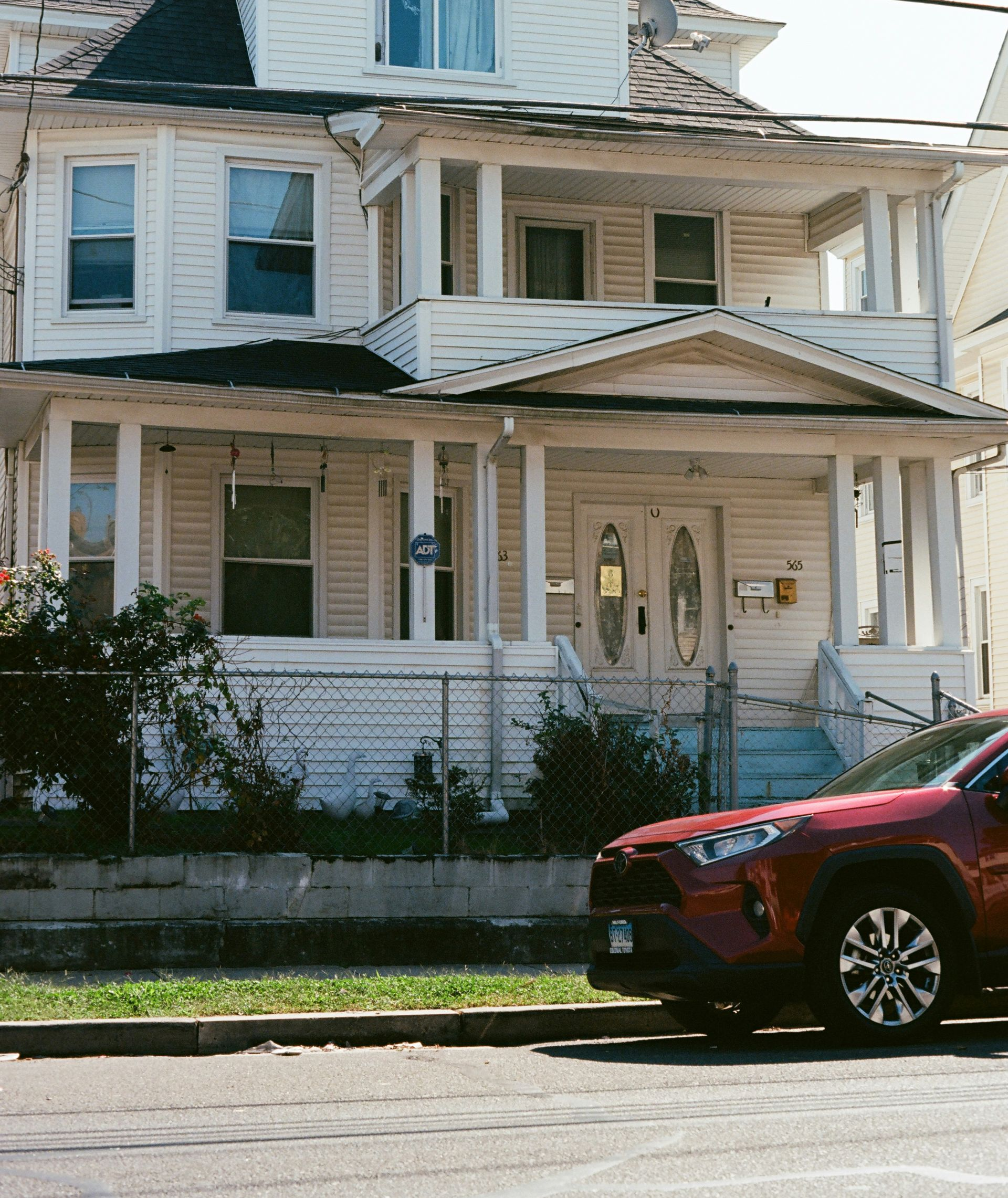A weathered, white, two-story house with a front porch, chain-link fence, and a red car parked in front.