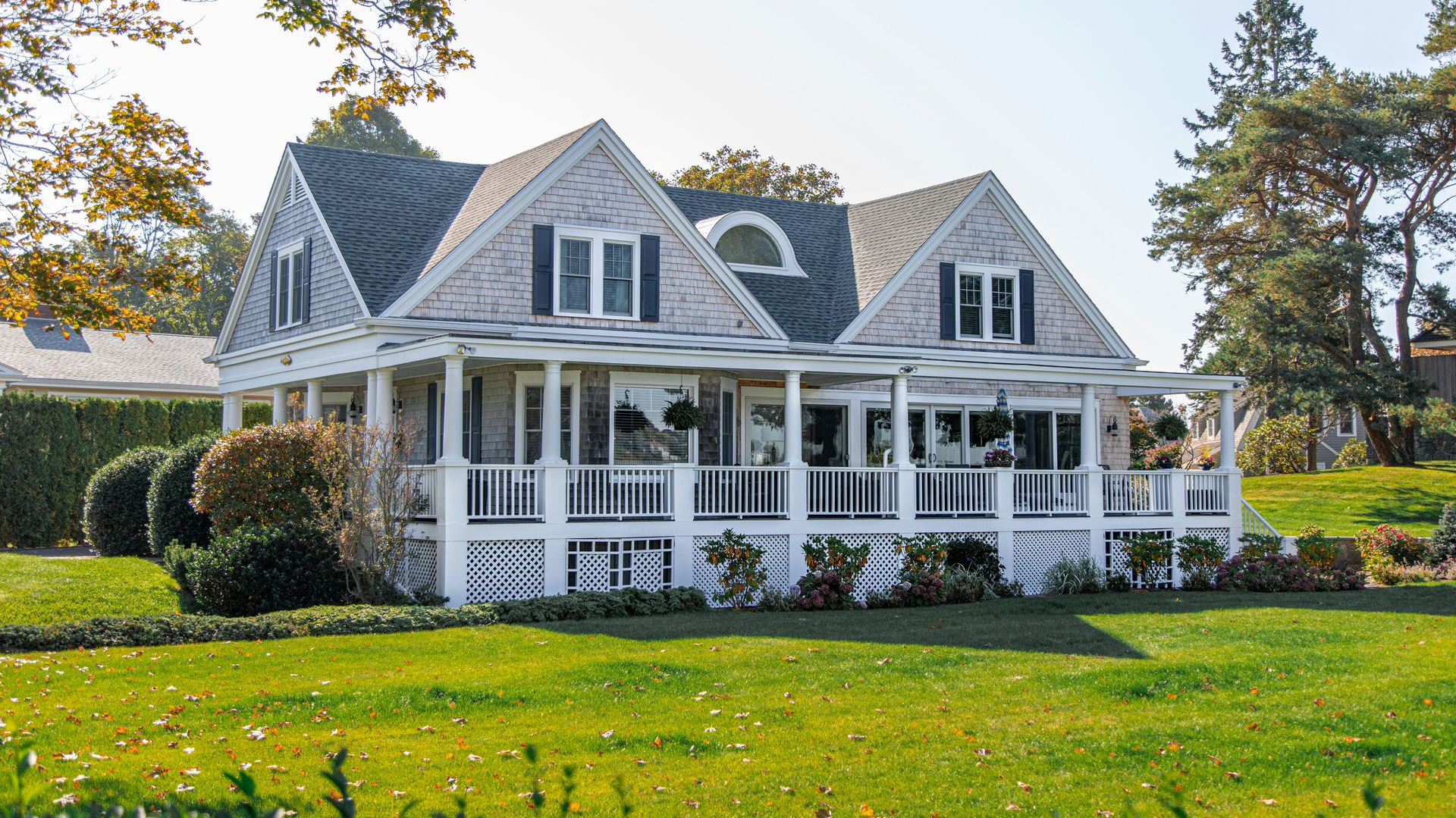 A gray shingled house with white trim and a wrap-around porch stands in a sunny, green lawn during the daytime.
