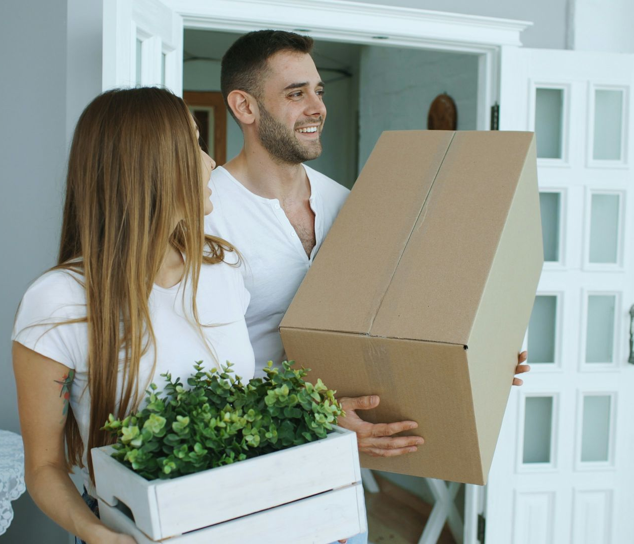 A couple stands in a doorway carrying a cardboard box and a potted plant, looking excited while moving into a new home.