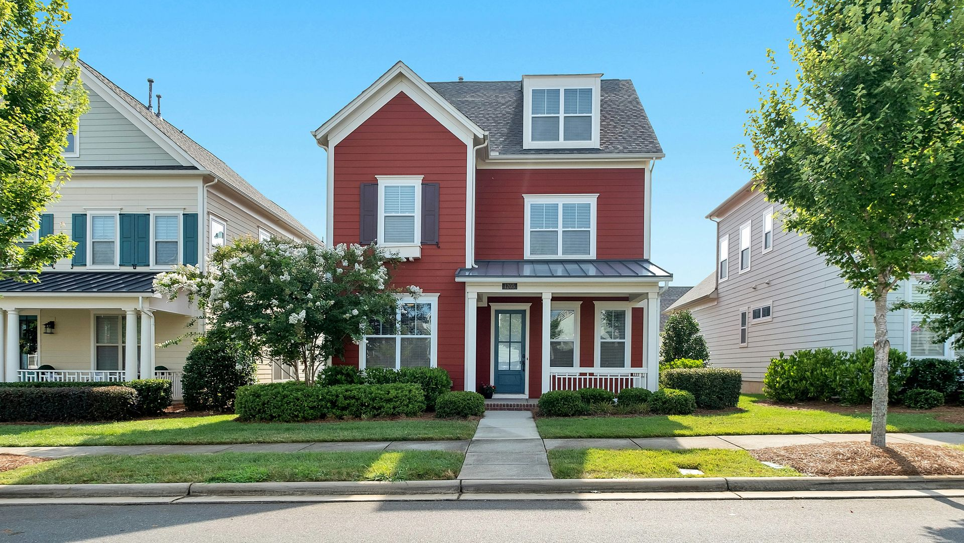 Two-story red house with white trim and porch; situated between similar houses with green lawns.