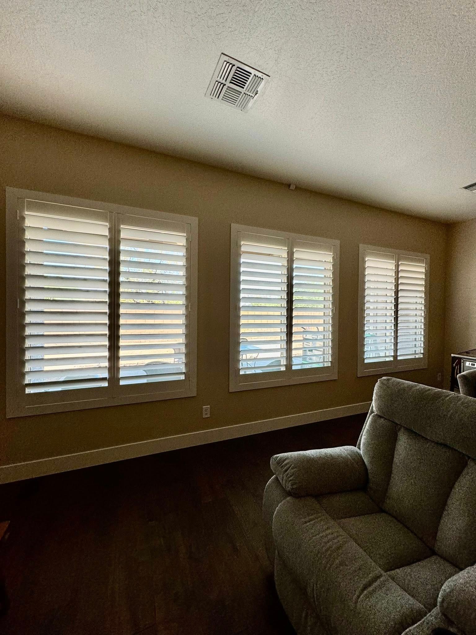 Three shuttered windows on a beige wall above dark wood flooring, and a gray armchair.