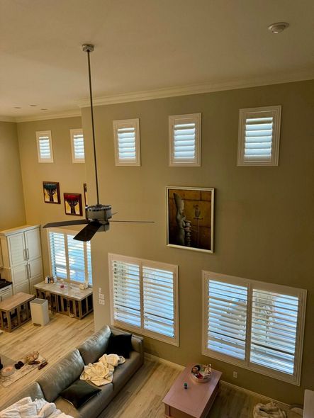 High-ceiling living room with windows and a couch; neutral walls, shuttered windows, ceiling fan.