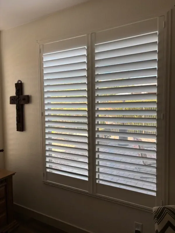 Two white plantation shutters on a window. A wooden cross hangs on the wall.