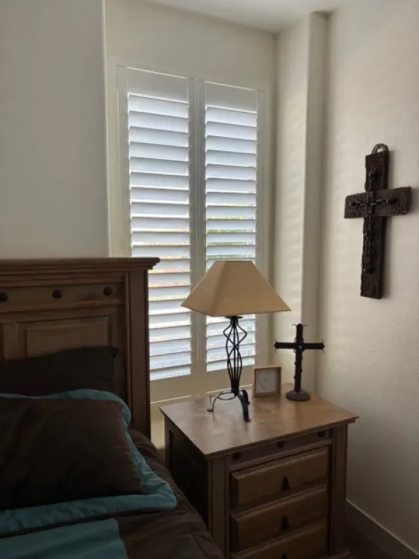 Bedroom corner with white shutters, nightstand with lamp, wooden cross on the wall.