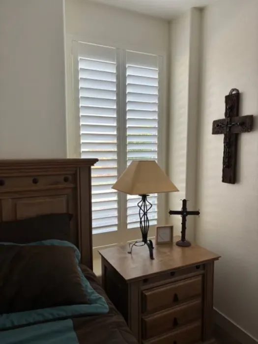 Bedroom corner with white shutters, nightstand with lamp, wooden cross on the wall.
