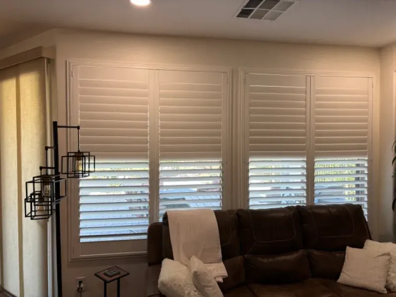Living room with white shutters on three windows above a brown leather couch.