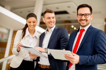 Three smiling business professionals in suits hold papers and a tablet in an office setting.