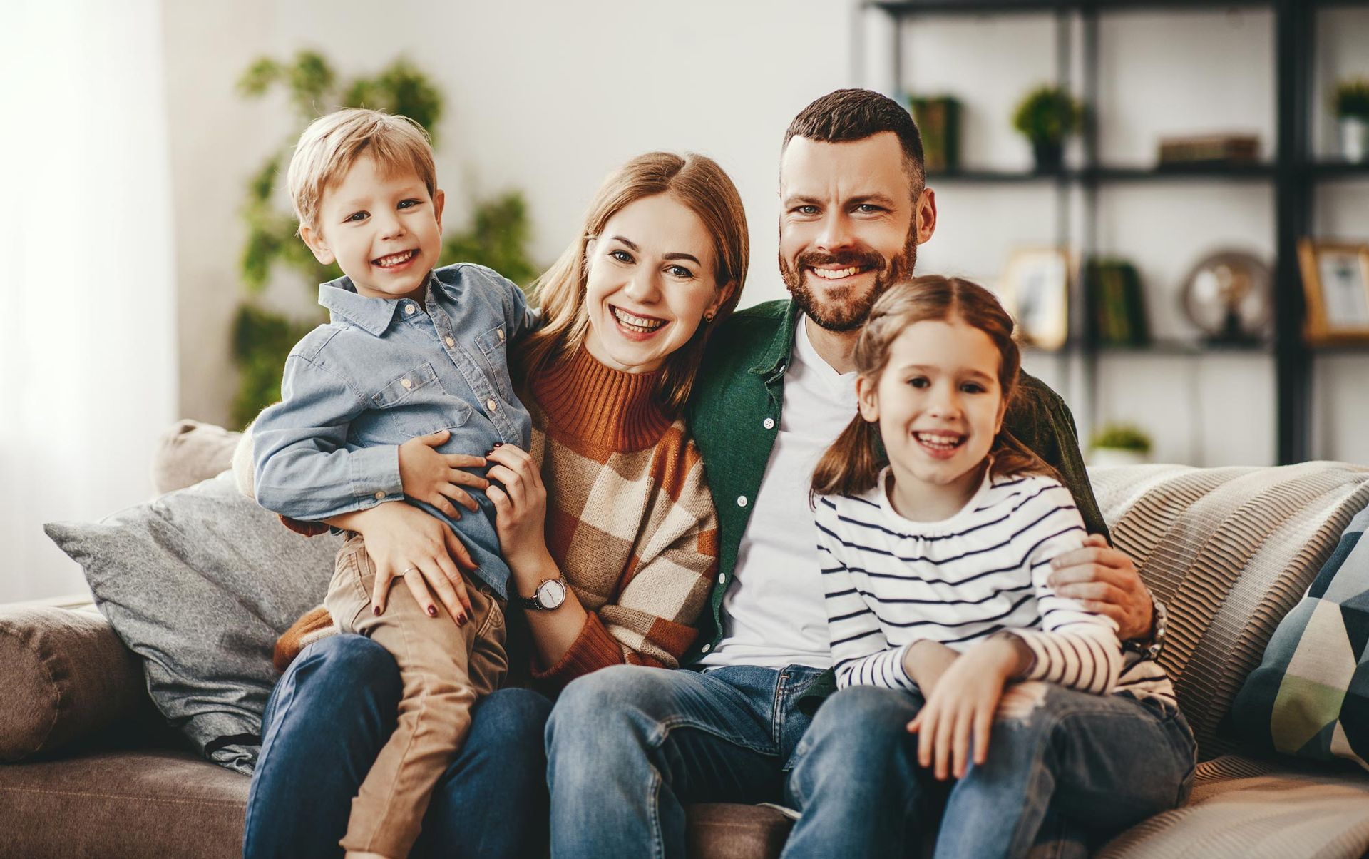 A smiling family of four sits close together on a couch. They are in a brightly lit living room.