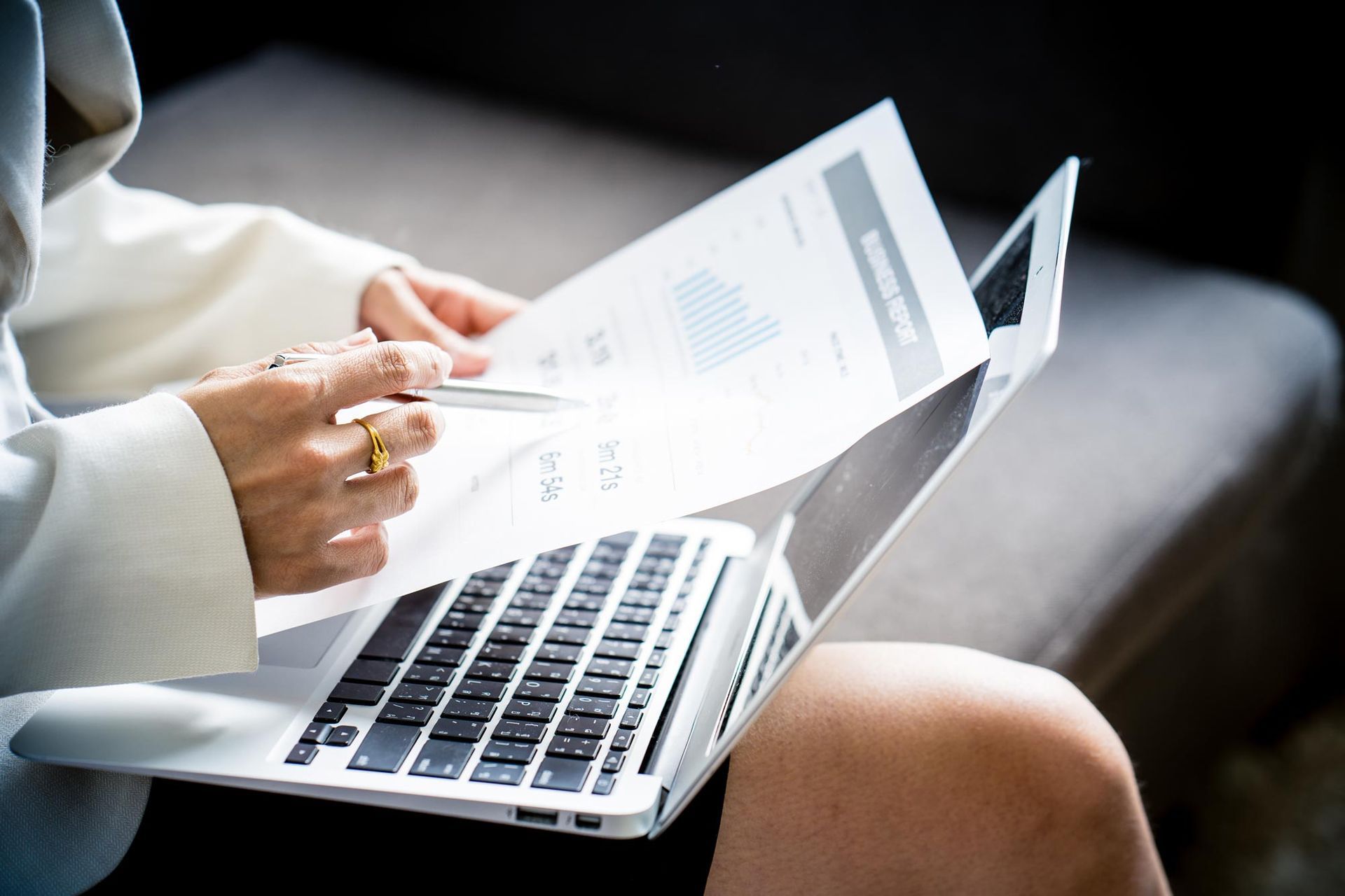 Person reviewing documents, holding a pen, and using a laptop on a couch.