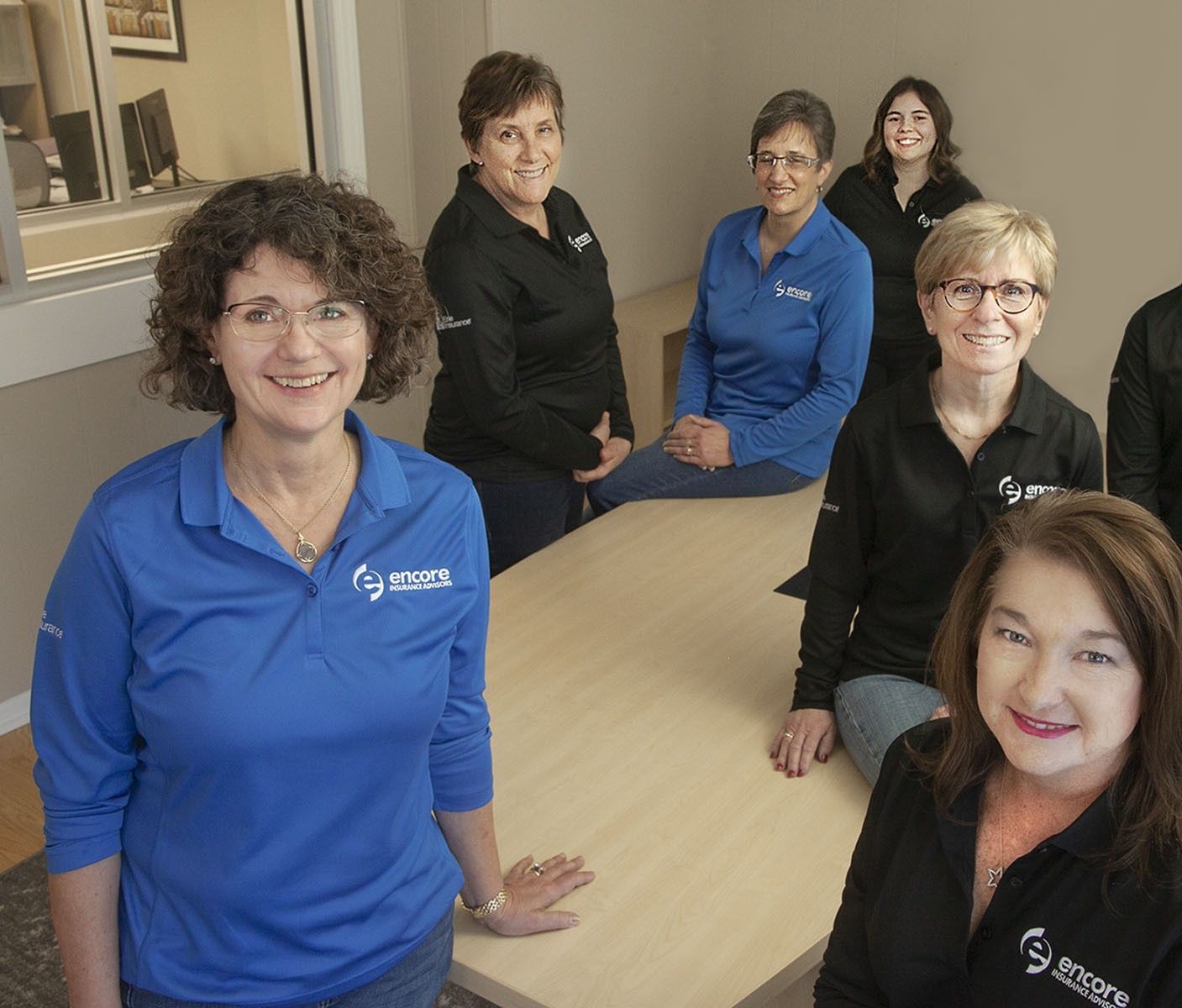 Group of seven women in blue and black polo shirts, smiling, in an office setting, gathered around a light-colored table.