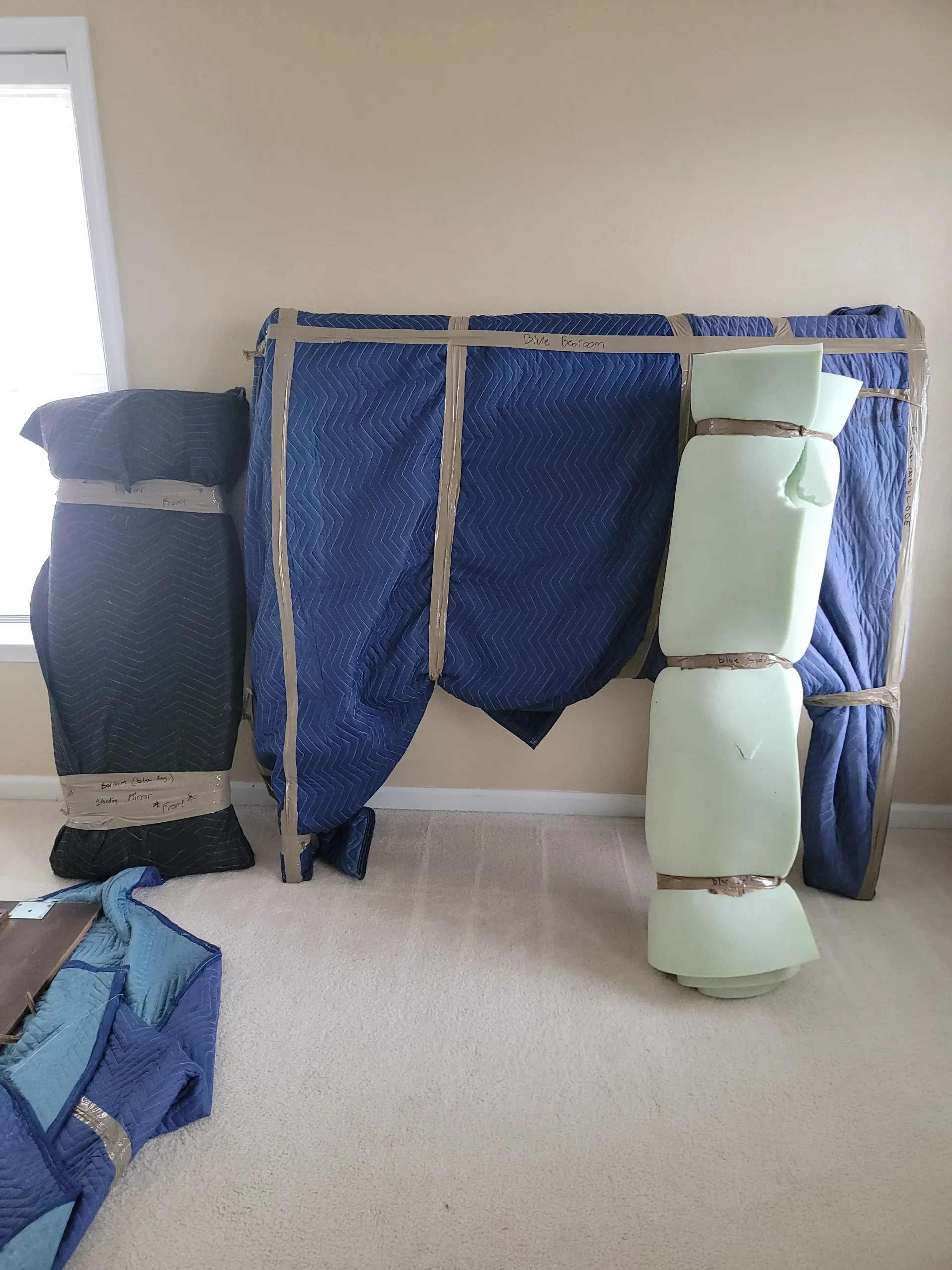 Large furniture pieces wrapped in blue moving blankets and packing foam, standing on a carpeted floor in a room.