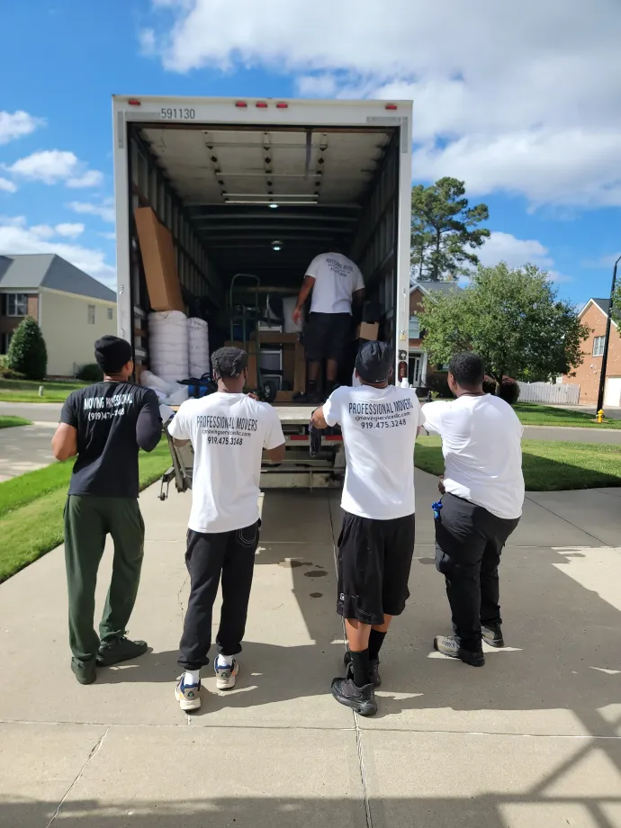 Five movers in uniforms load equipment into the back of a parked moving truck on a sunny residential street.