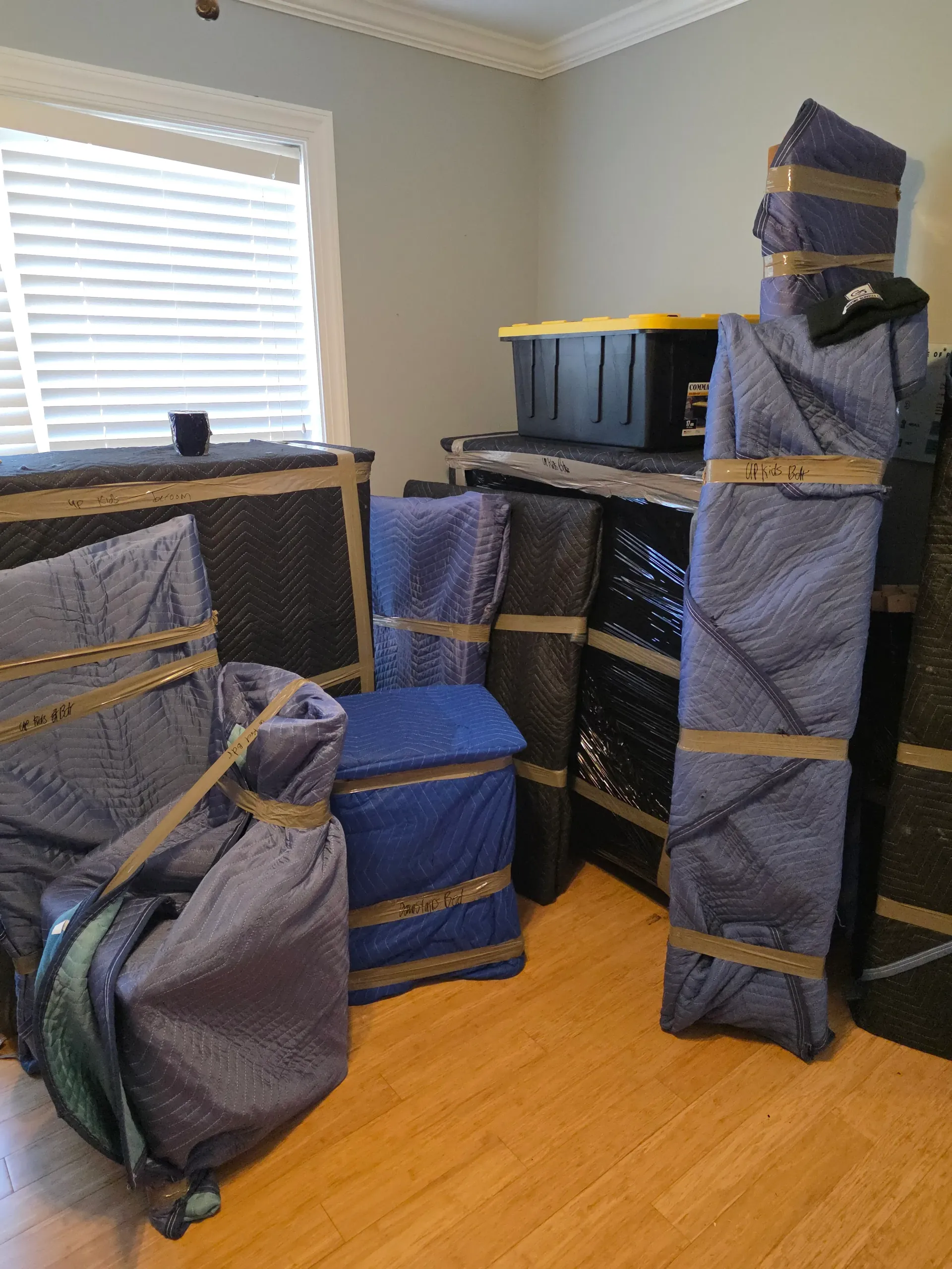 A room filled with various items wrapped in blue moving blankets and secured with tape, including boxes and furniture.