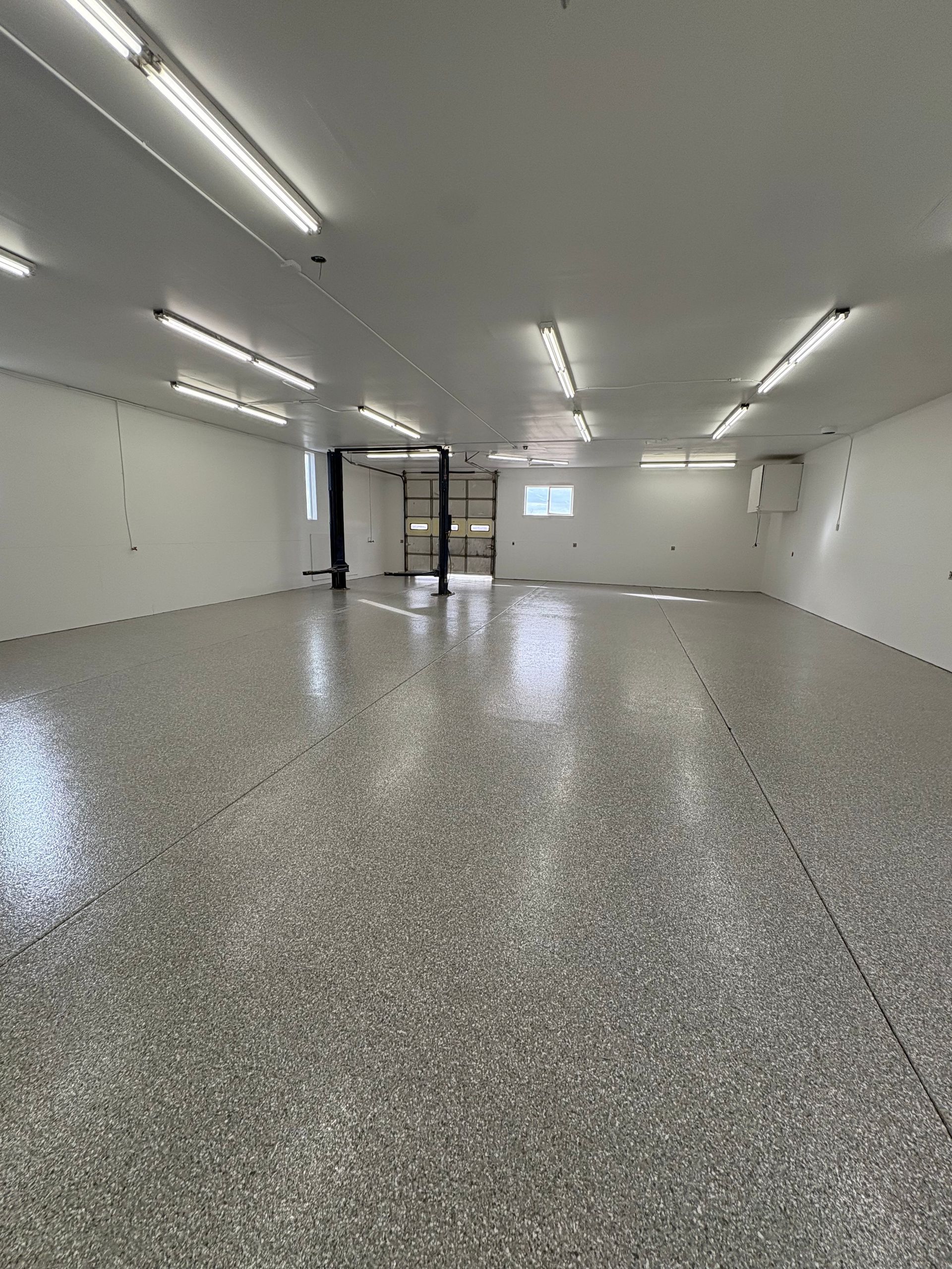 A wide, empty room with white walls, a glossy gray speckled floor, and bright fluorescent ceiling lights.
