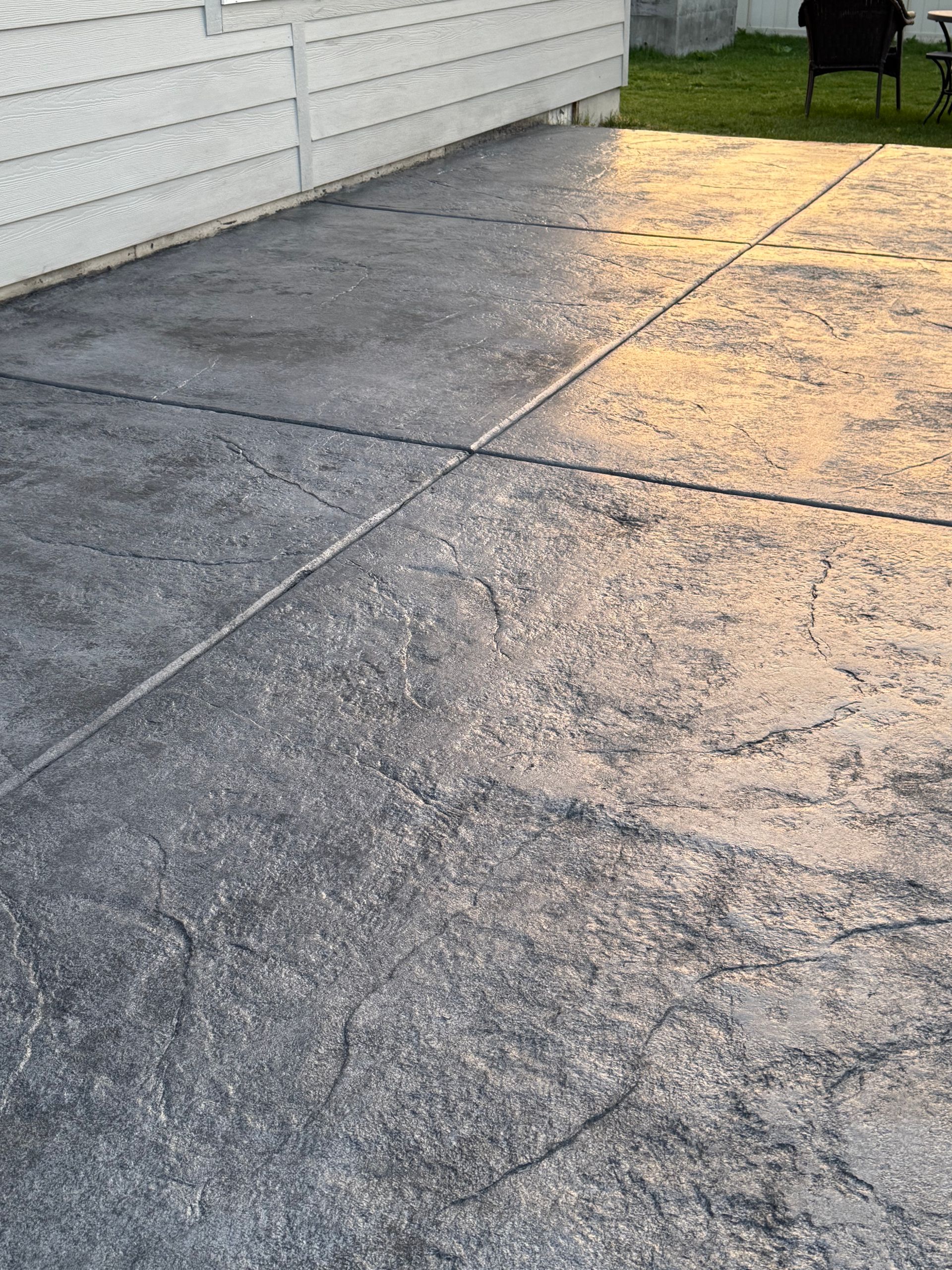 A close-up of a gray stamped concrete patio with square patterns, bordered by the white siding of a house at sunset.