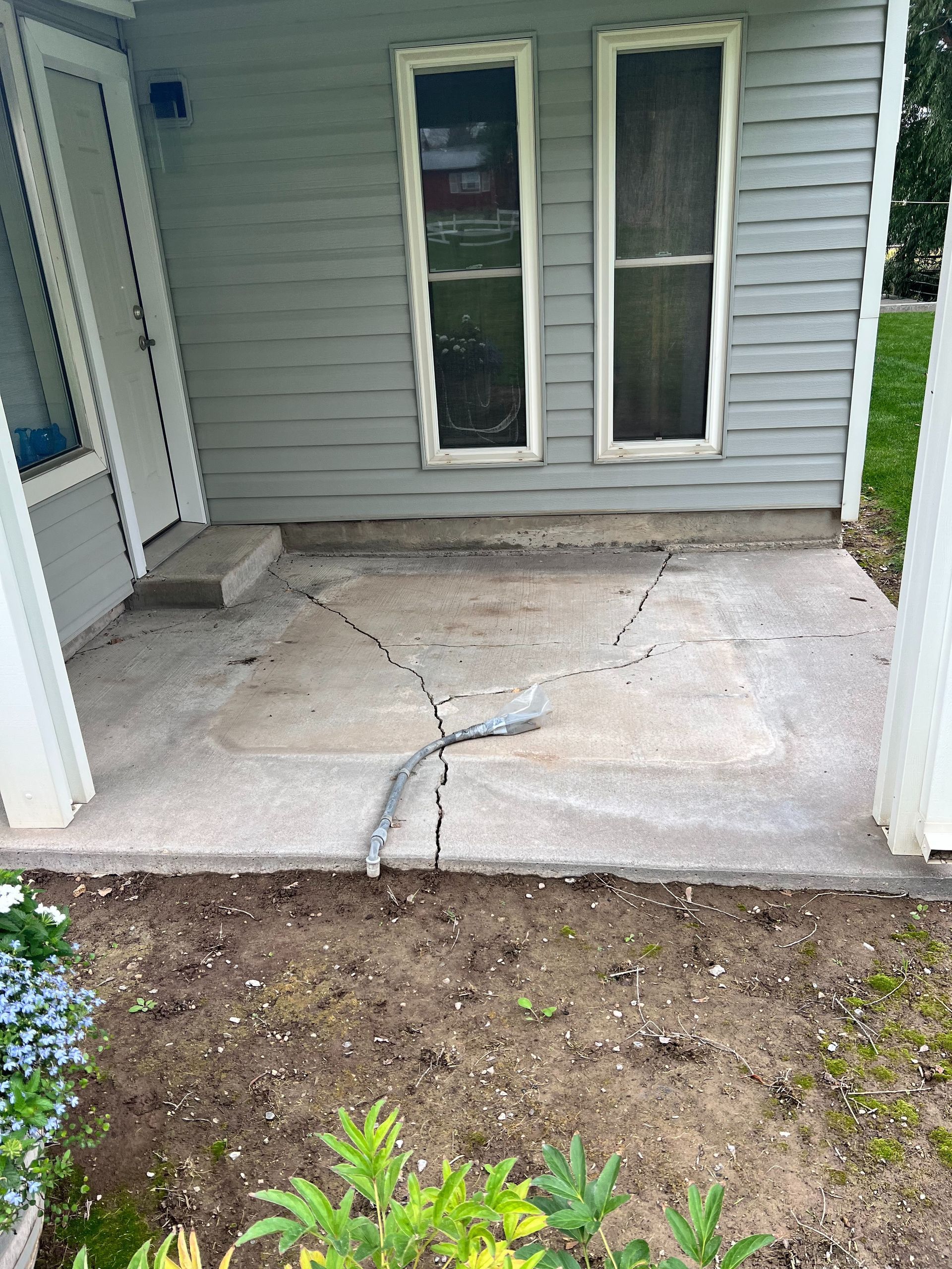 A concrete porch area in front of a house with grey siding, showing significant cracks and damaged pavement.