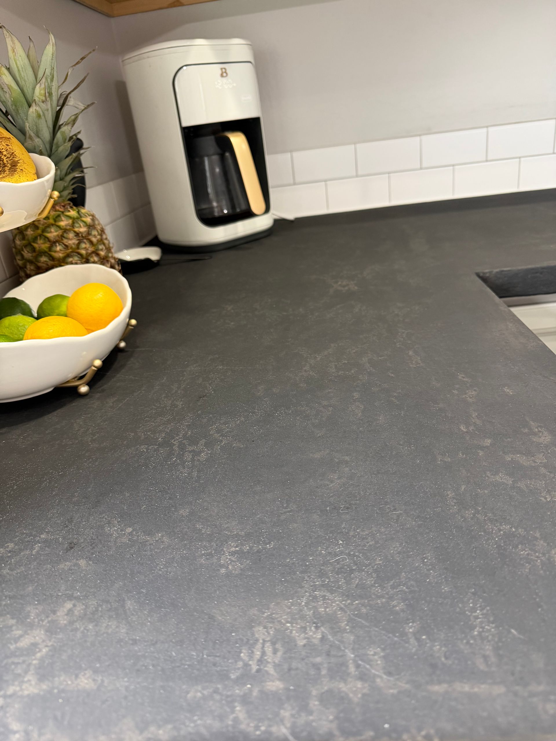 A white coffee maker sits on a dark gray kitchen counter next to a white bowl of lemons and a pineapple.