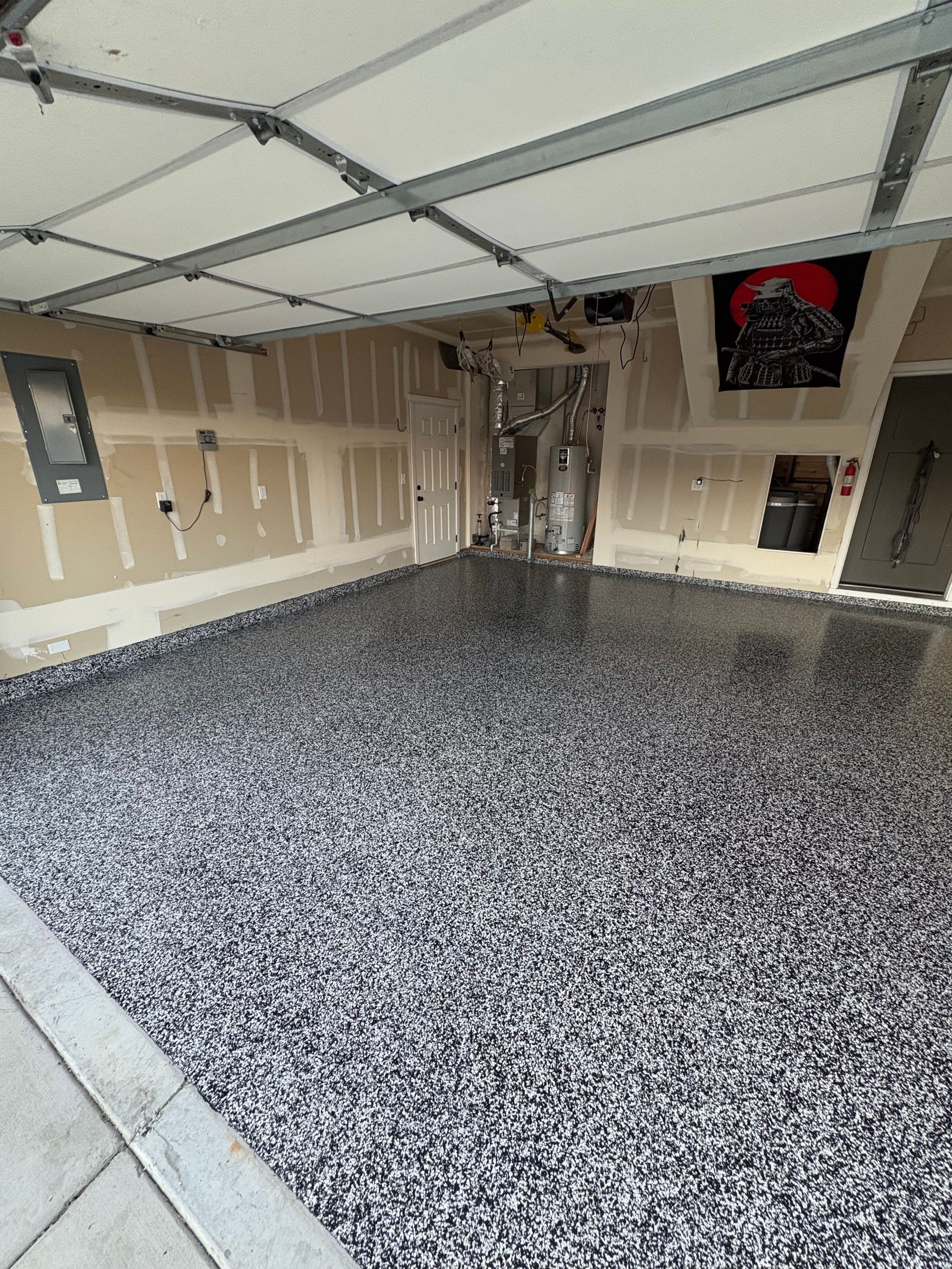 A garage with a newly applied gray and black speckled epoxy floor, unfinished drywall, and exposed ceiling beams.