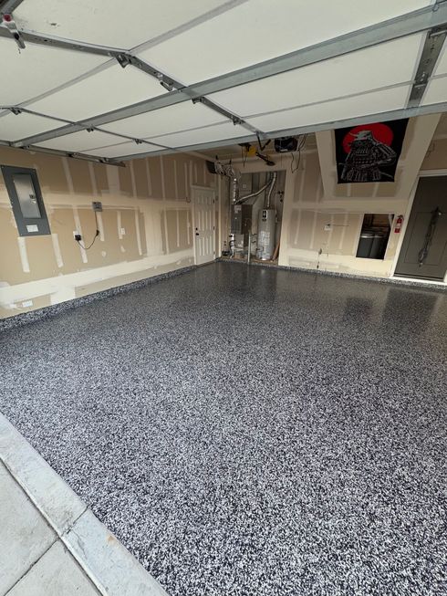 A garage with a newly applied gray and black speckled epoxy floor, unfinished drywall, and exposed ceiling beams.