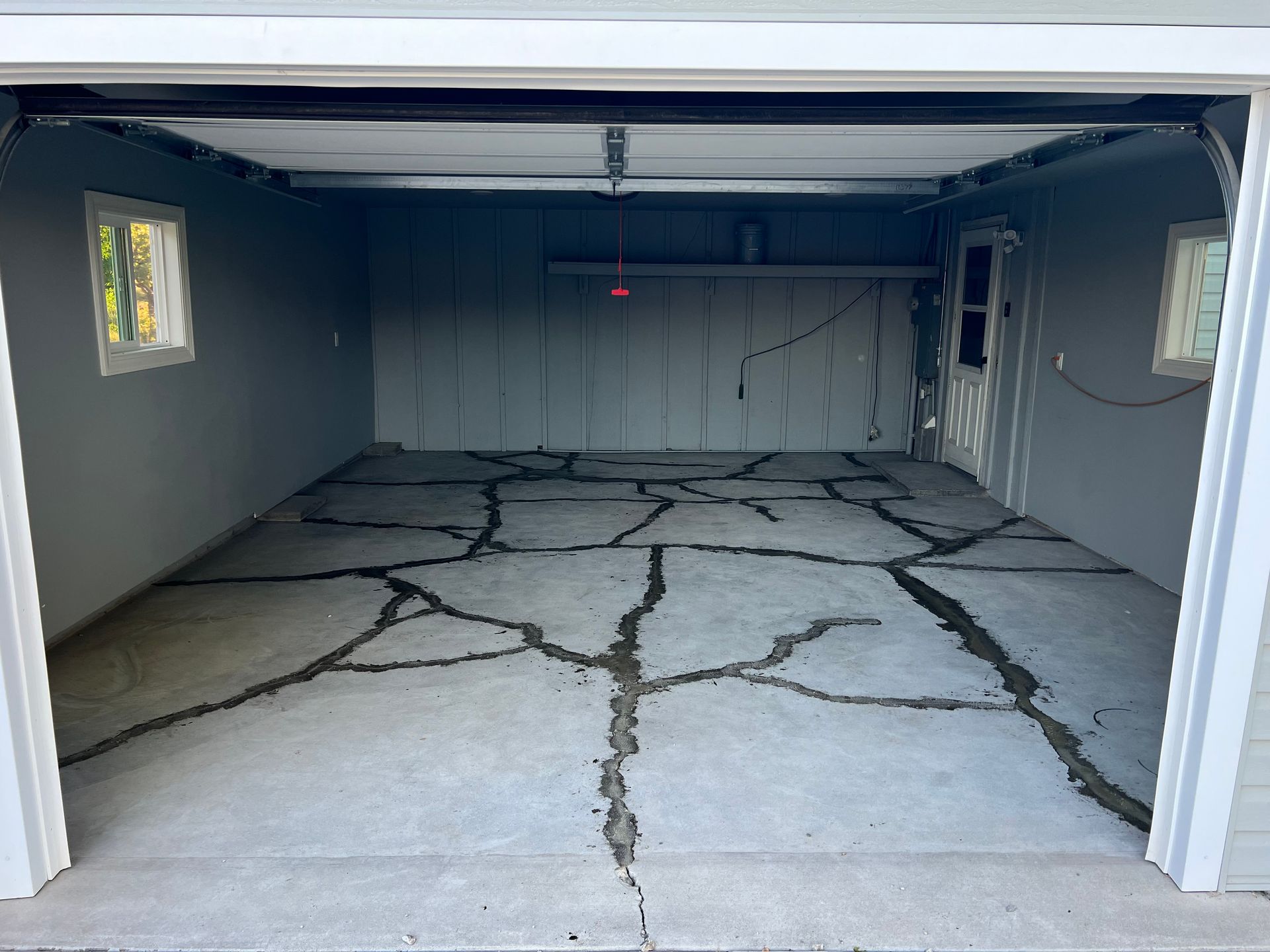 A gray garage interior with a concrete floor featuring extensive, wide, dark-filled cracks spanning the surface.