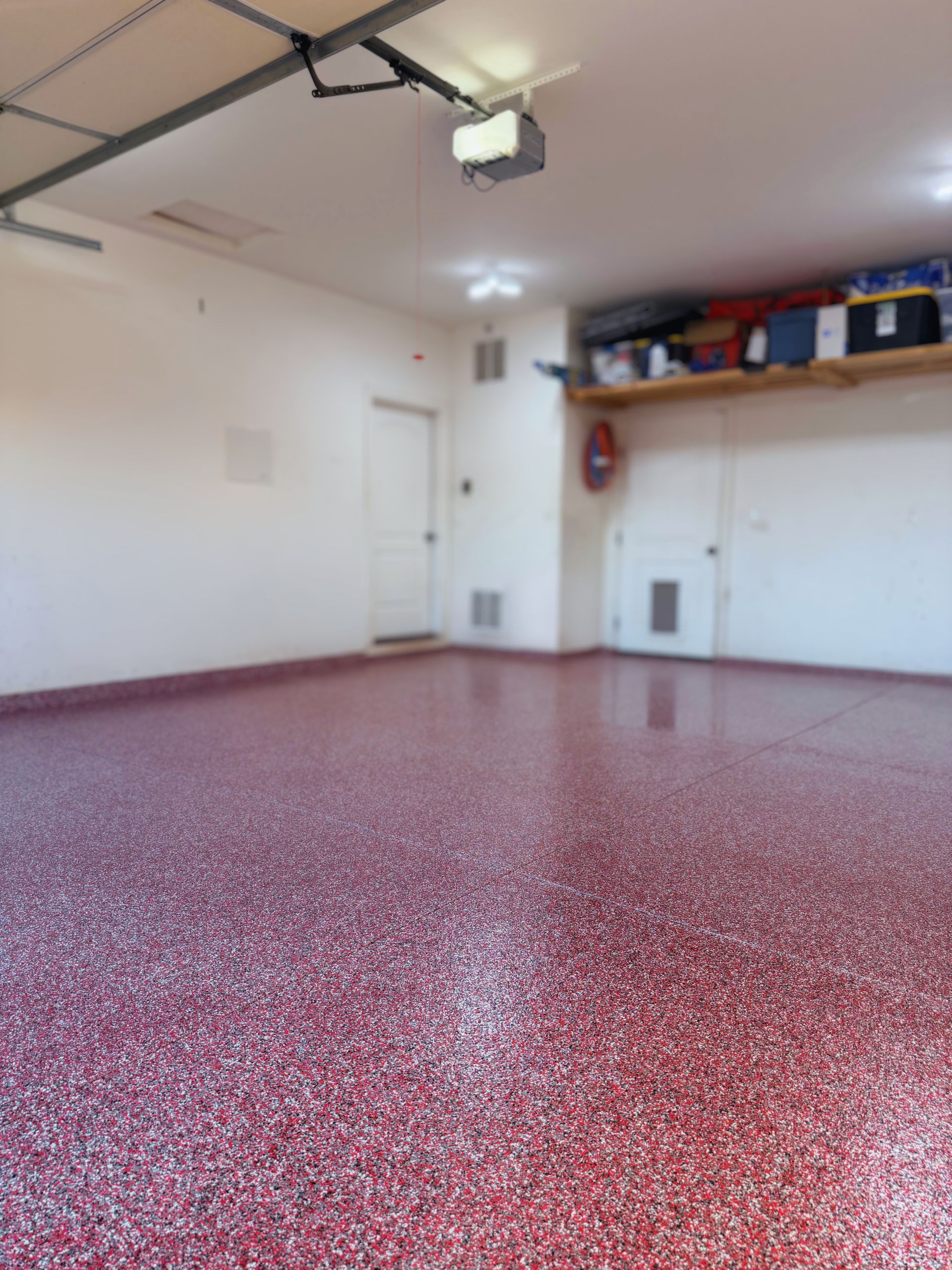 An empty garage interior with a glossy, red and white speckled epoxy floor, white walls, and overhead storage shelving.
